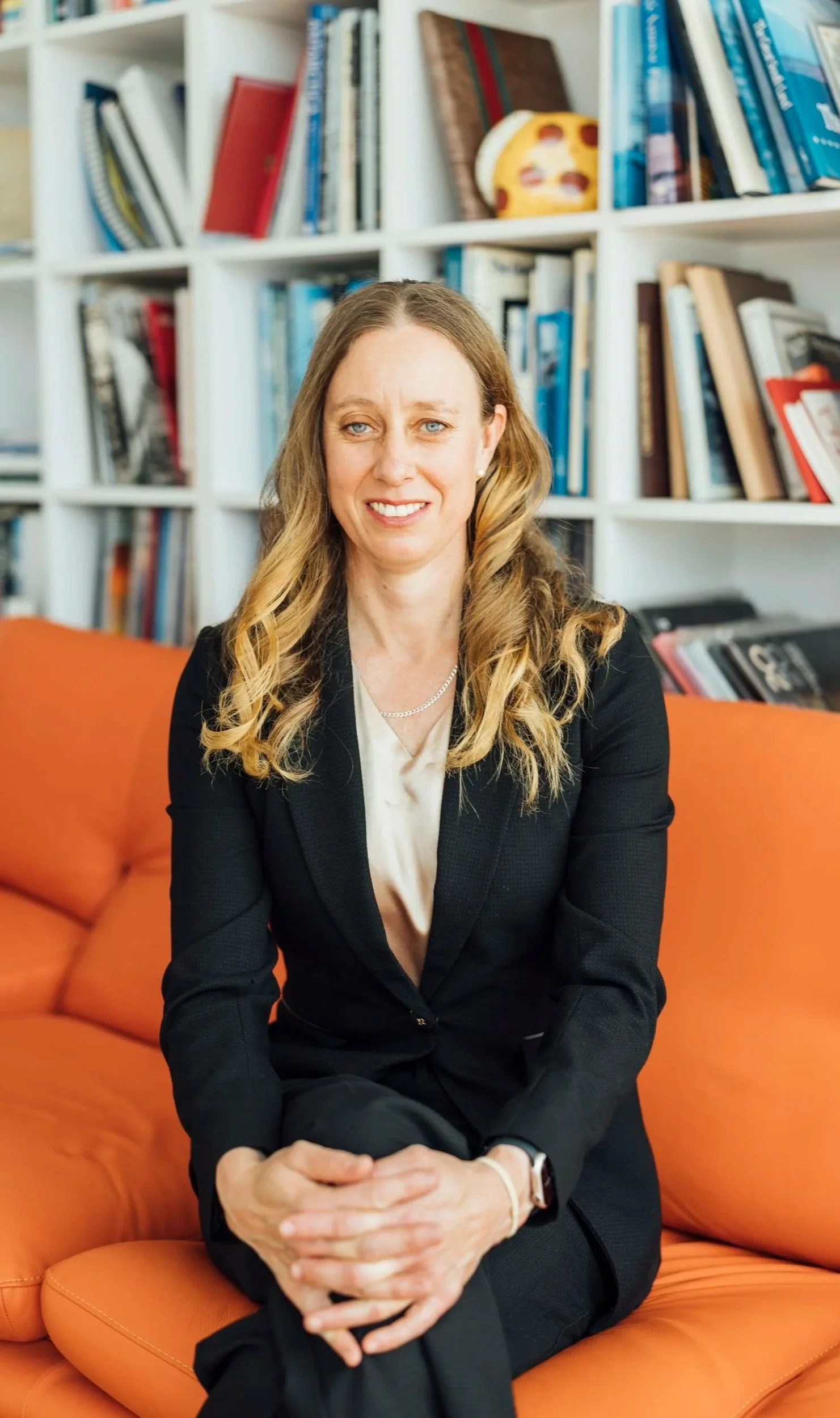 A female orthopaedic surgeon with long blonde hair, wearing a black blazer and cream blouse, sitting on an orange sofa in front of a bookshelf filled with books.