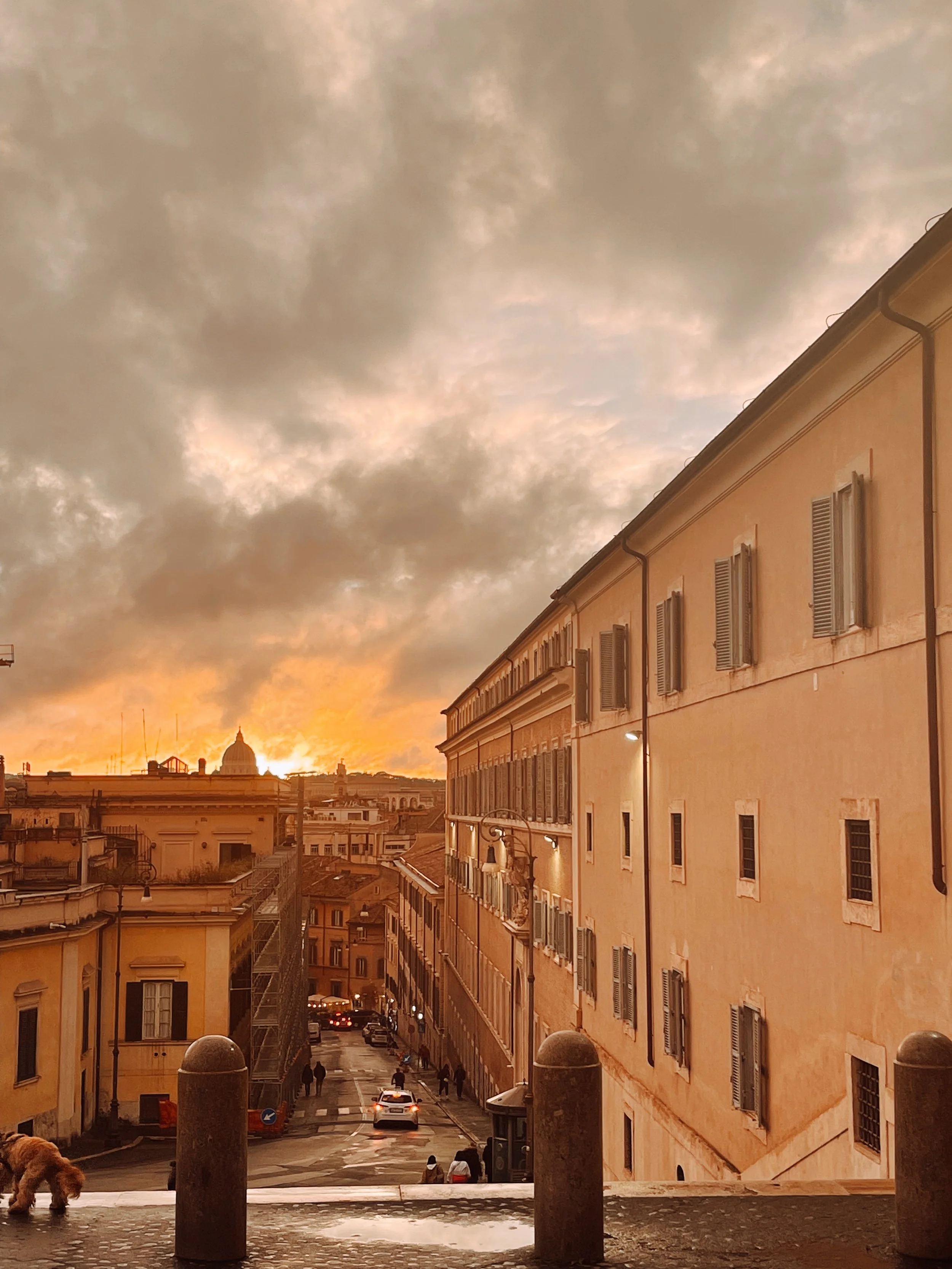 View of a city street at sunset with cloudy sky, buildings on either side, and people walking on the sidewalks.