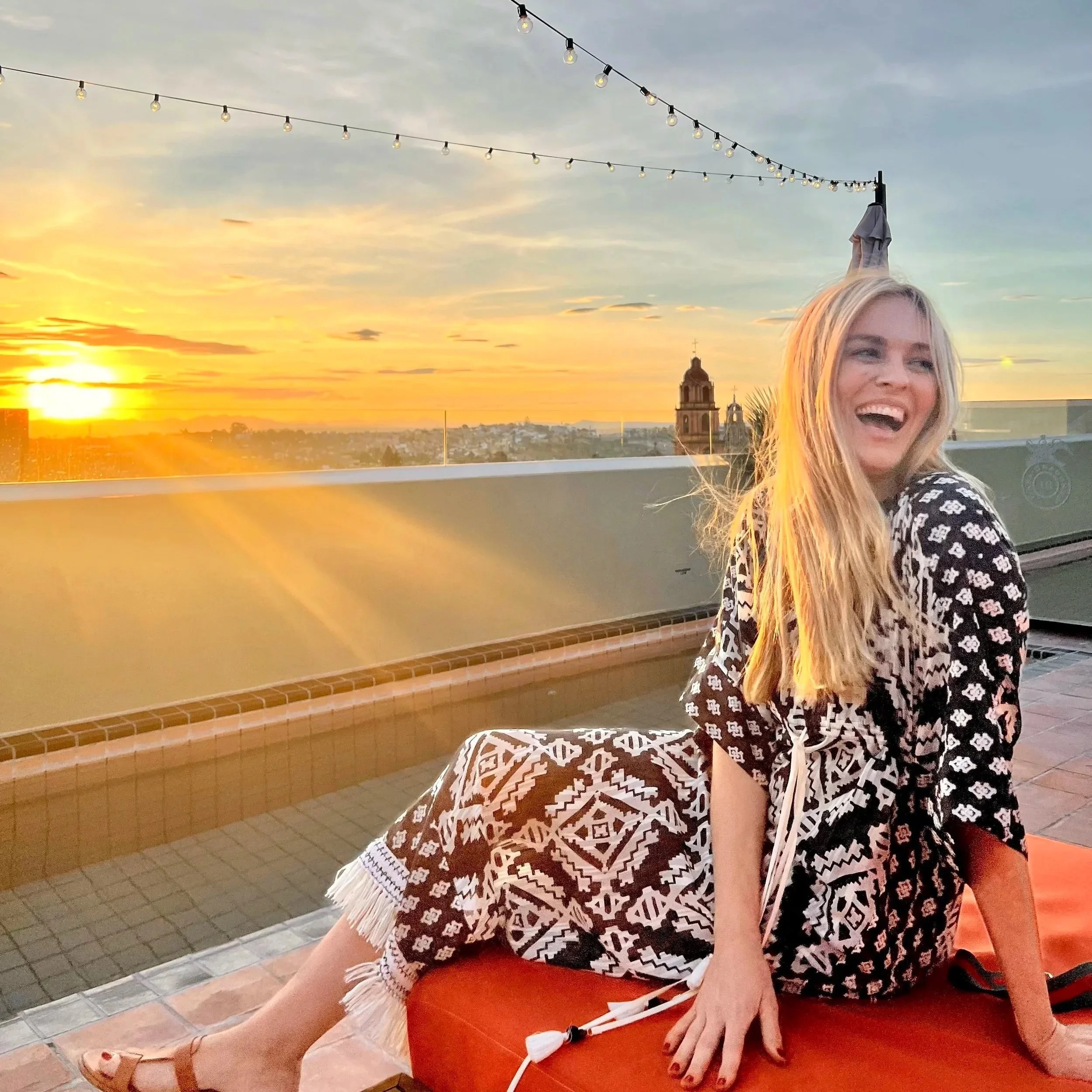 Young woman sitting on a lounge chair, smiling, with sunset and cityscape in the background, outdoor setting with string lights overhead.