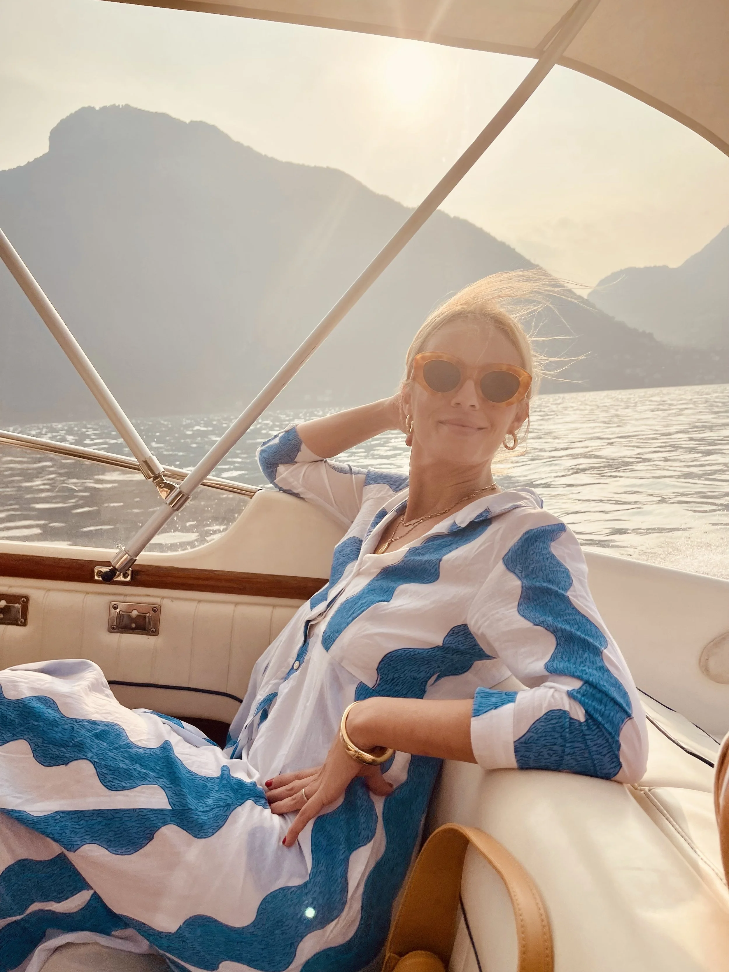 Woman with blonde hair and large sunglasses relaxing on a boat during sunset over mountains and water.