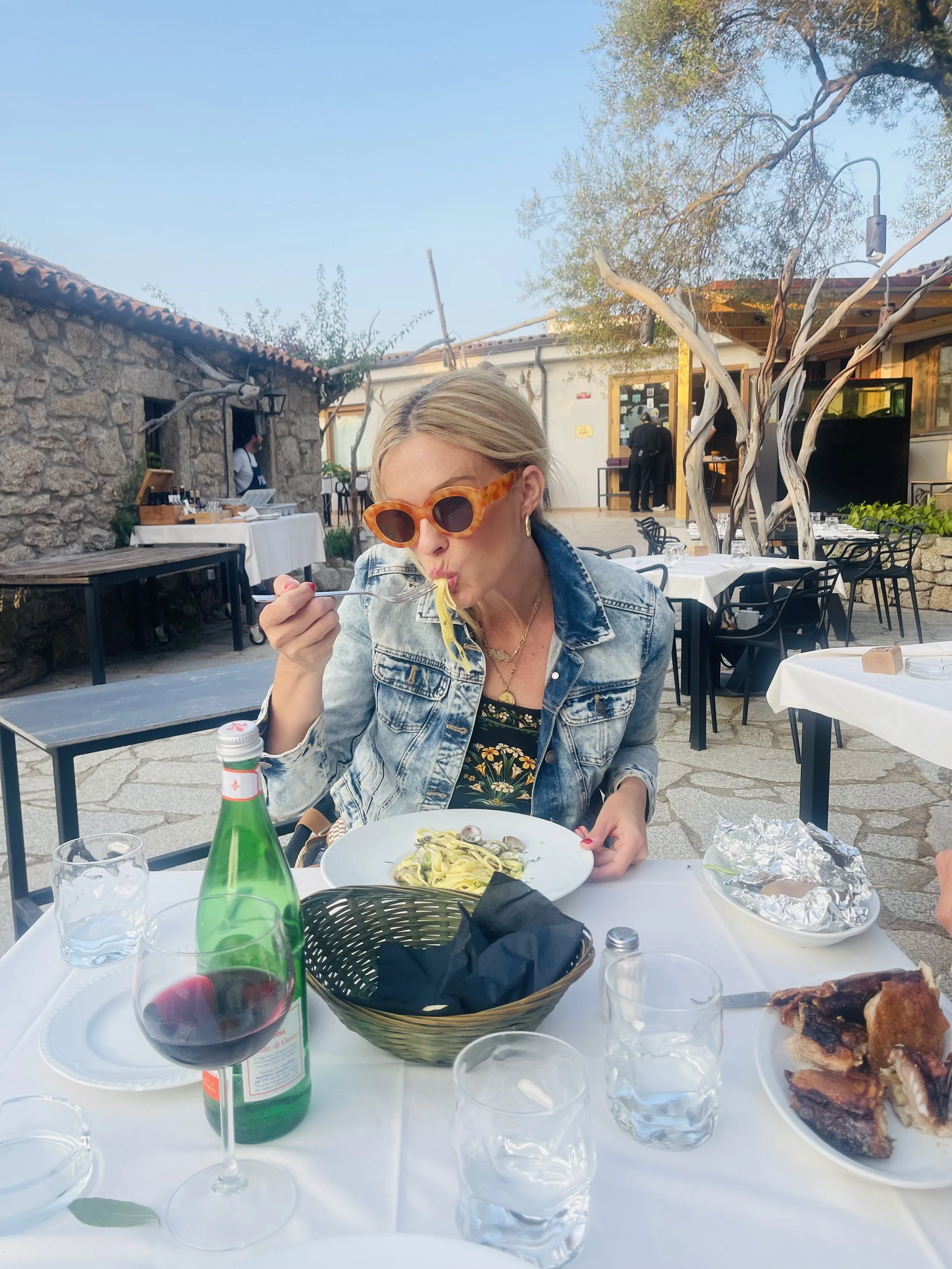 A woman with blonde hair and tortoise shell sunglasses eating pasta at an outdoor restaurant table with a glass of red wine and plates of food.
