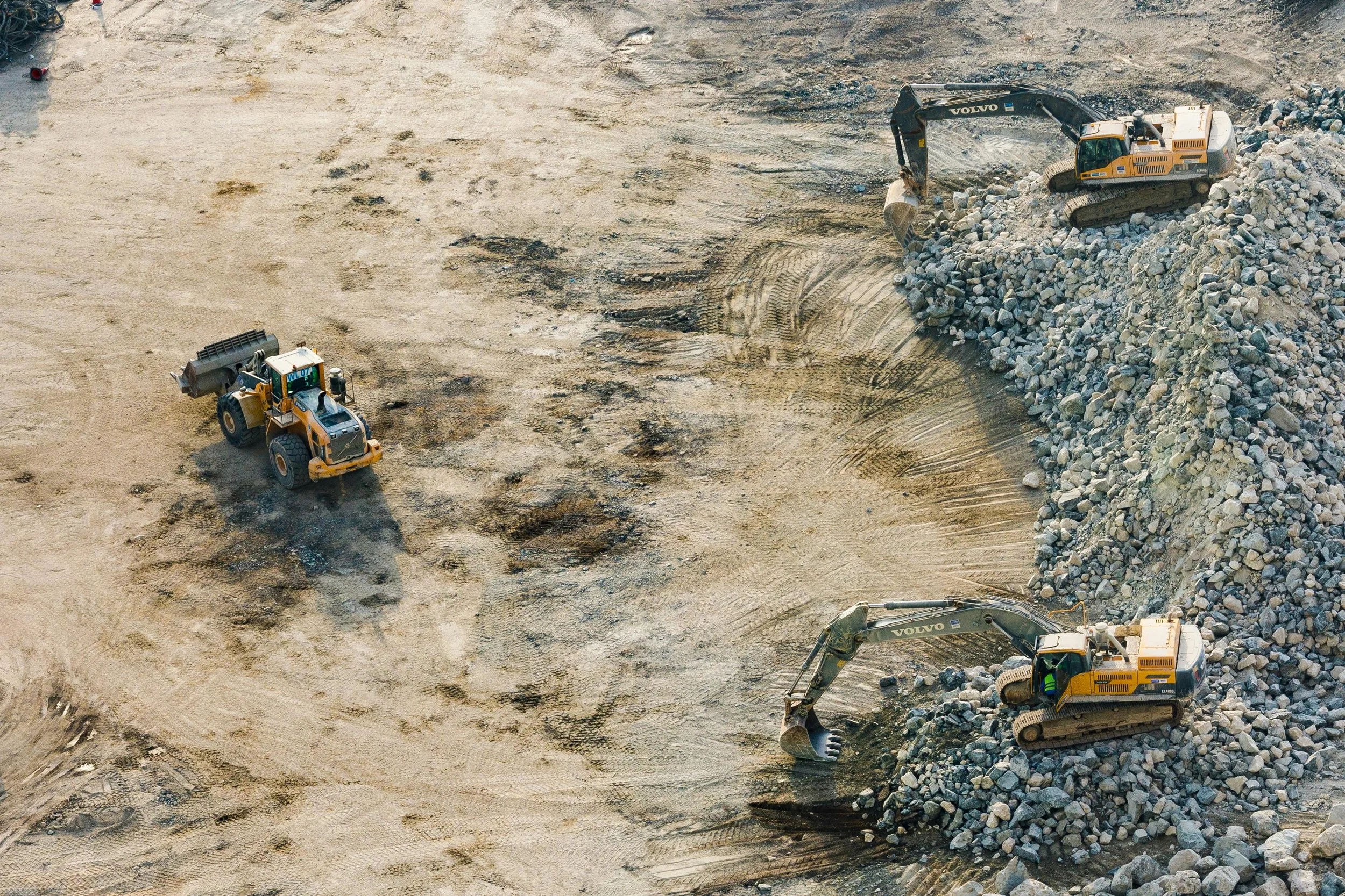 Three construction excavators working on a rocky, dirt-covered site. Two are on a pile of rocks, and one is clearing dirt with a bucket.