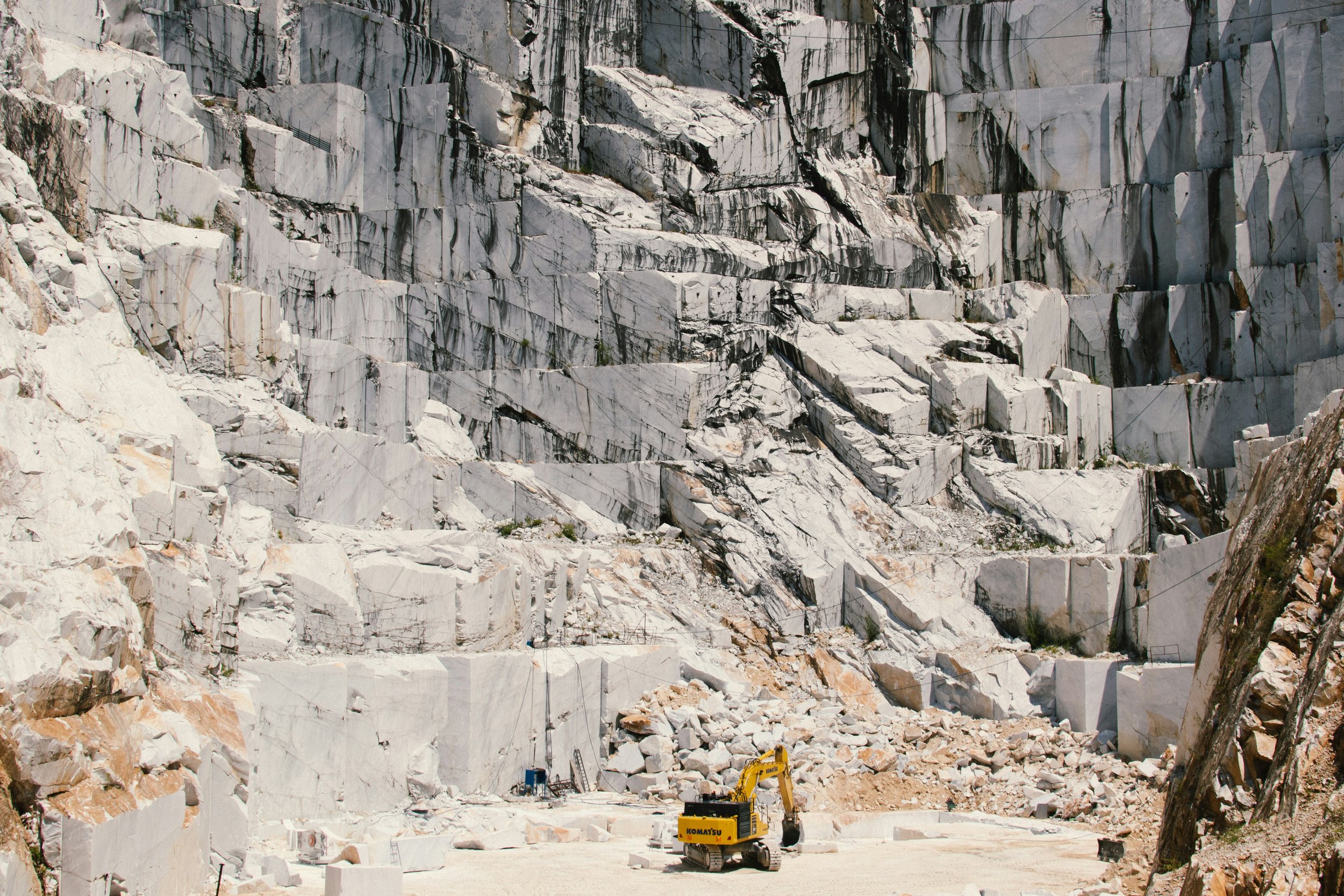 A large white marble quarry with a small yellow excavator at the bottom center, surrounded by stone rubble.