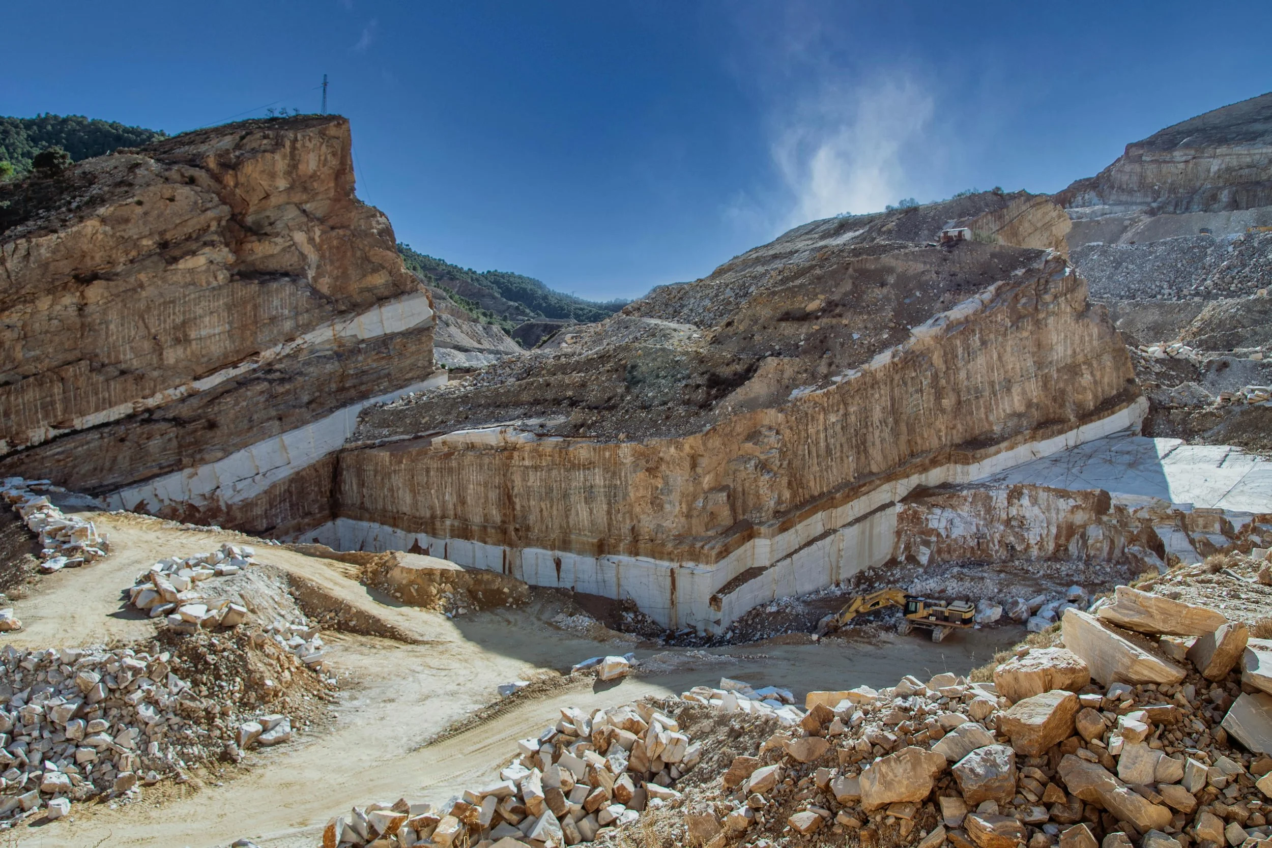 Large open-pit quarry with colorful layered rock formations, dirt roads, excavation equipment, and scattered rocks with a mountain and blue sky in the background.