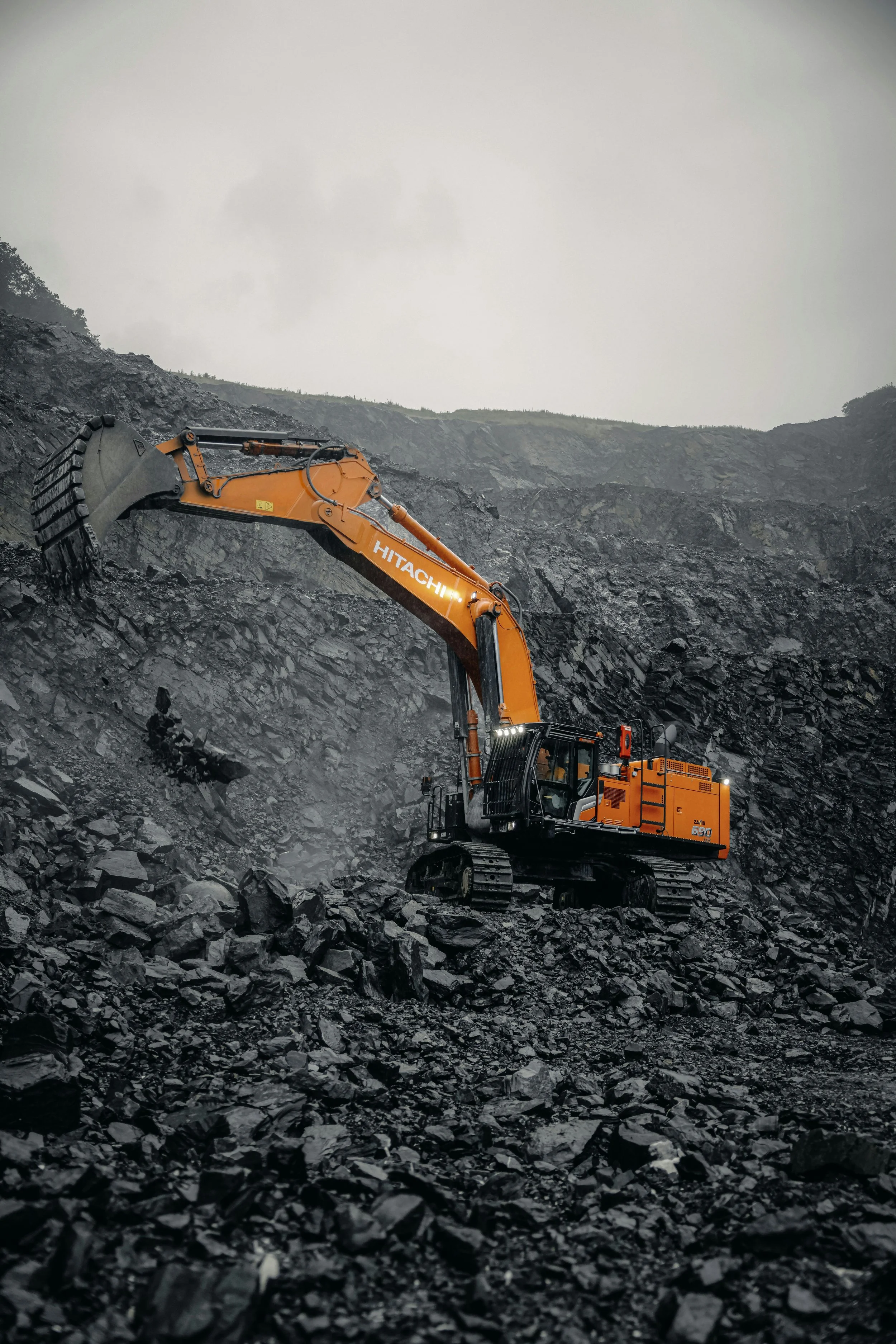 A large orange Hitachi excavator moving rocks on a black rocky landscape under an overcast sky.