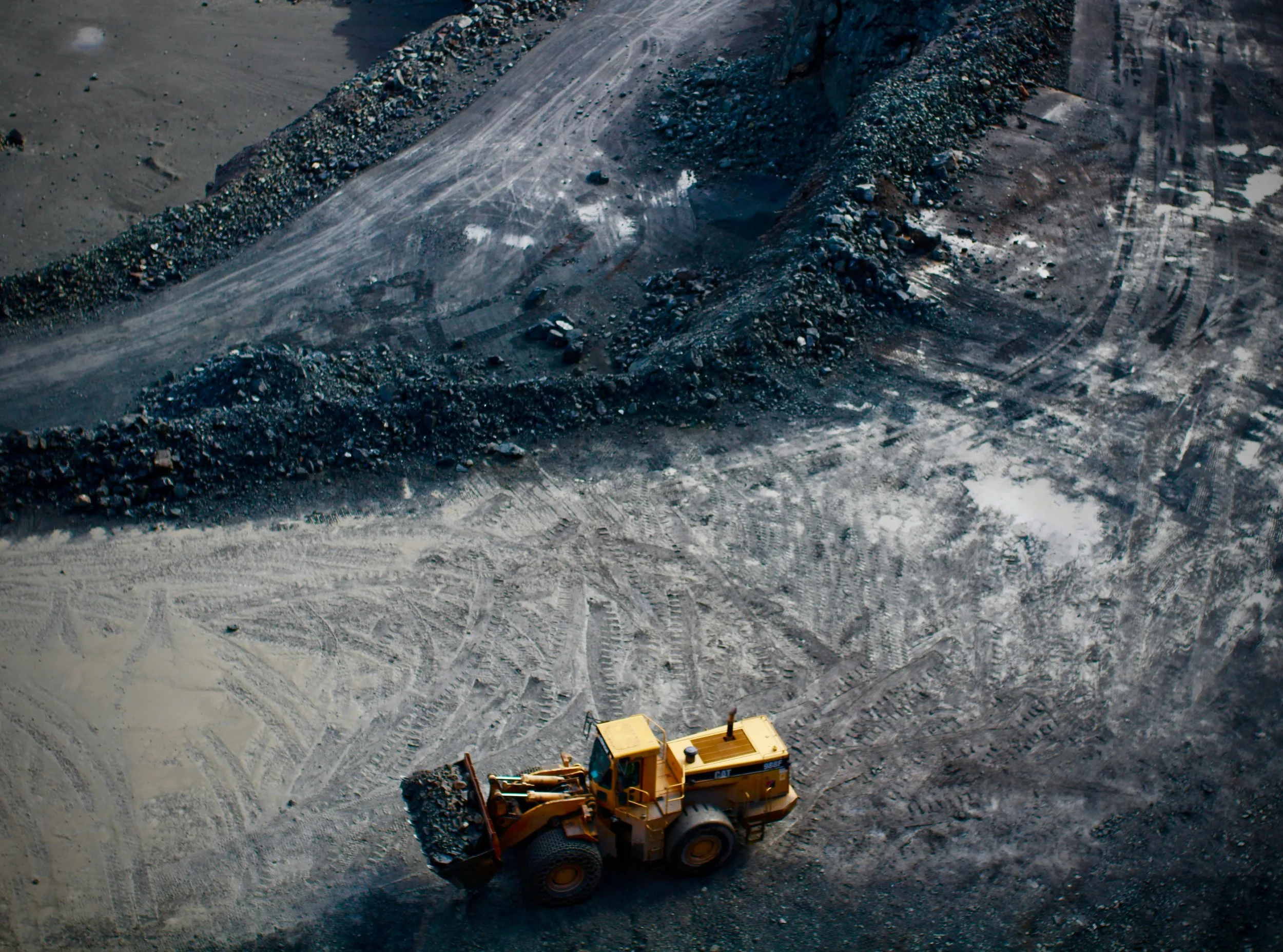 A yellow construction vehicle on a dirt lot with tire tracks, near a large pile of rocks and dirt at a mining or construction site.