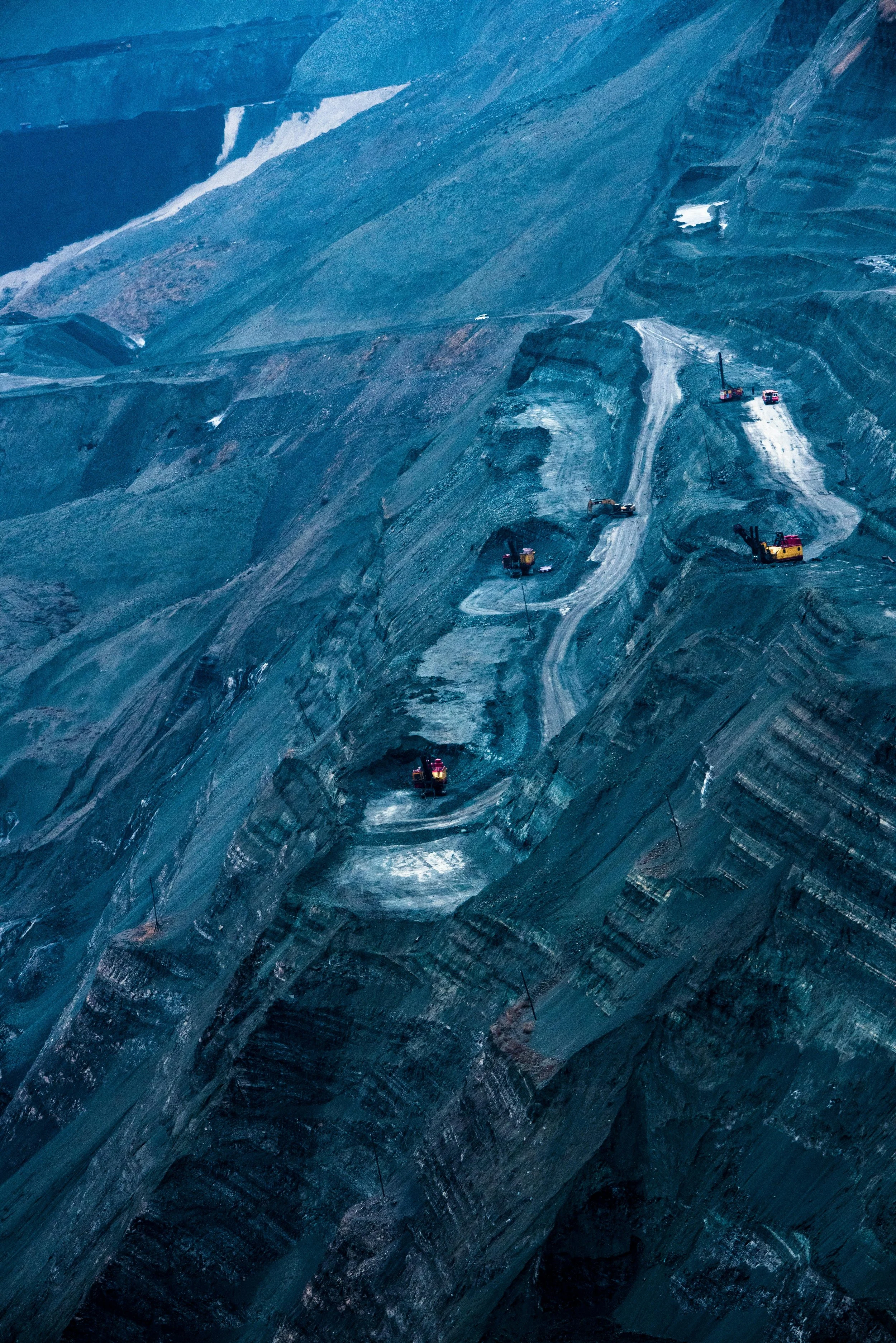 Construction vehicles working on a large open-pit mining site with terraced, layered rocky terrain.