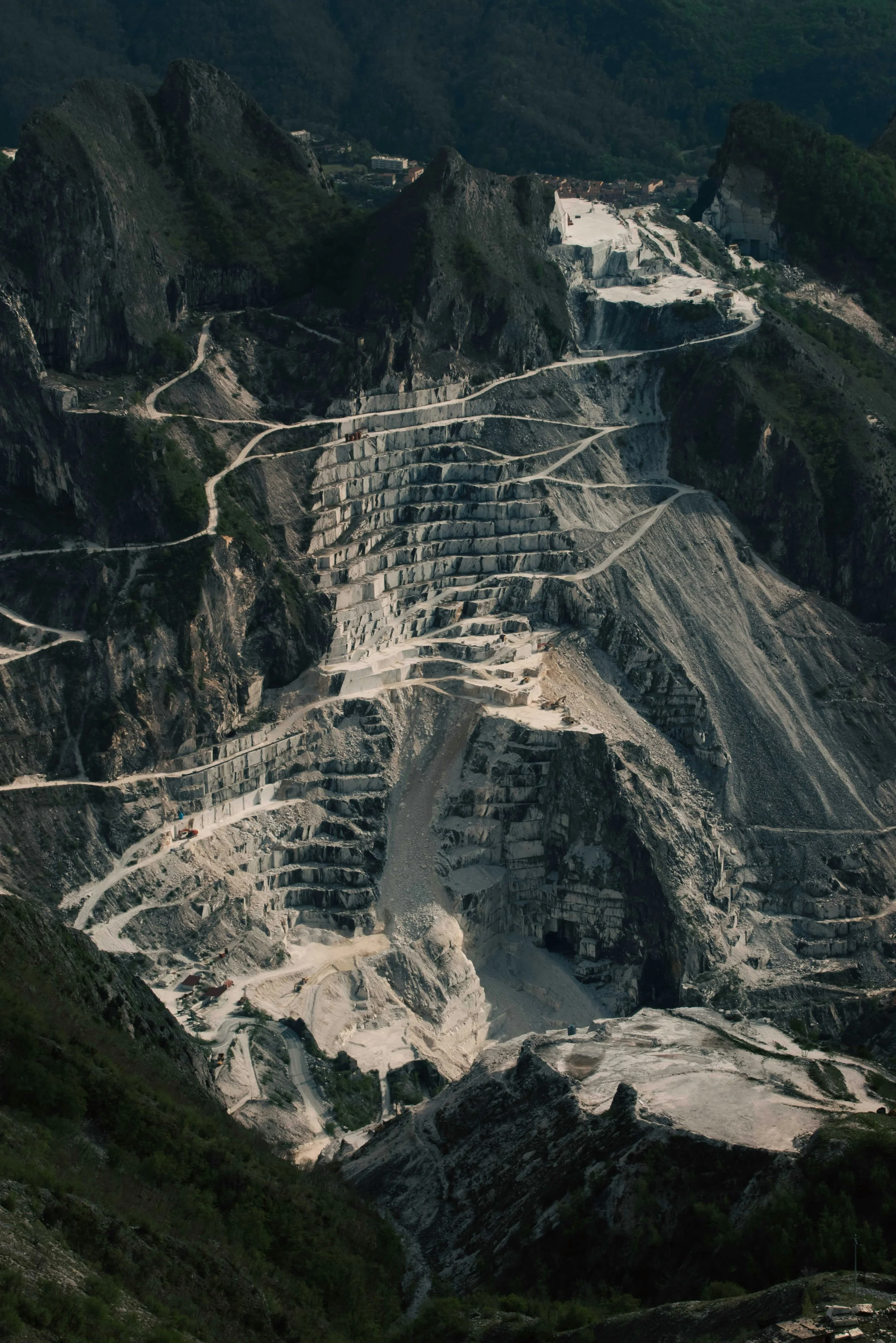 A mountainside with a large open-pit marble quarry, showing terraced steps carved into the mountain for extracting marble, with winding pathways around the site.