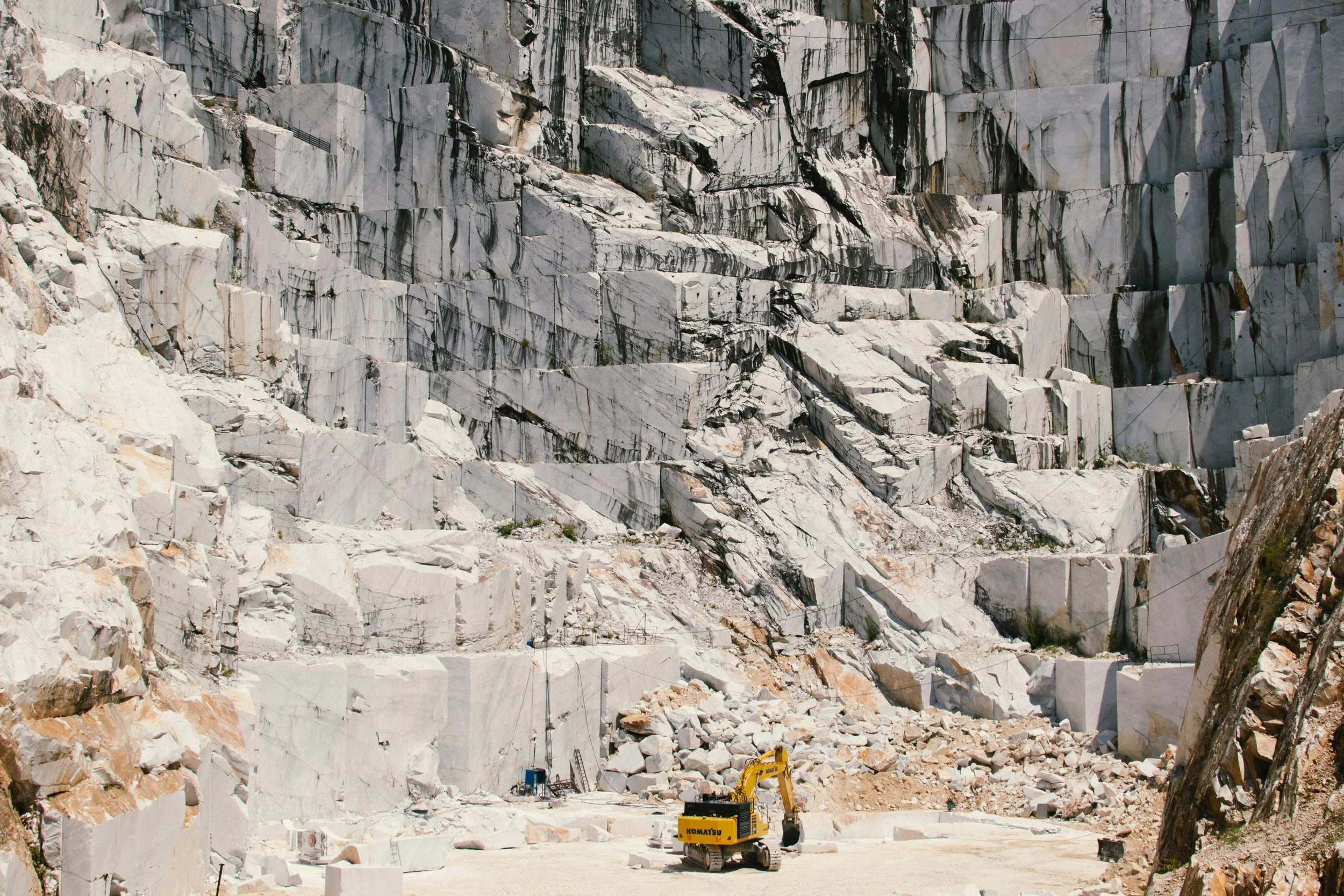 A large marble quarry with a yellow excavator in the foreground and massive white marble blocks and rough cuts on the rocky walls in the background.