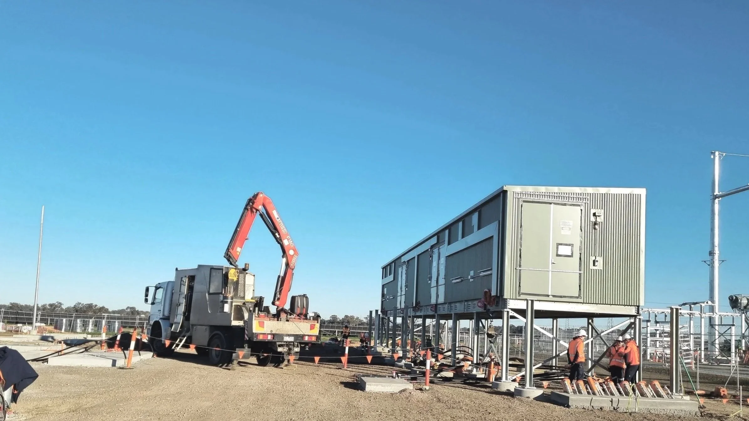Construction site (solar farm) with a winch truck and crane lifting equipment, workers in orange vests and helmets, and a large modular electrical building structure on stilts against a clear blue sky.