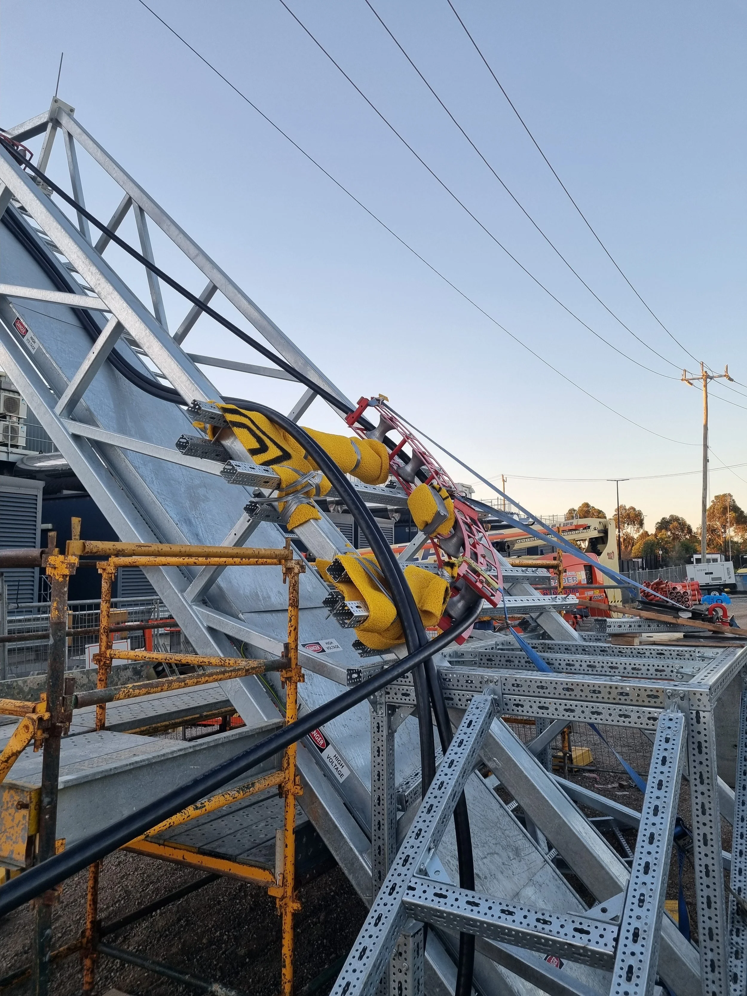 Construction site with metal scaffolding, yellow cushioned safety harnesses, power cables, and utility poles under a clear sky.