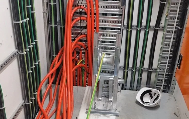 Electrical or network wiring setup with multiple orange and green cables along a metallic framework, a cable management strap, and a white protective helmet on a work surface.