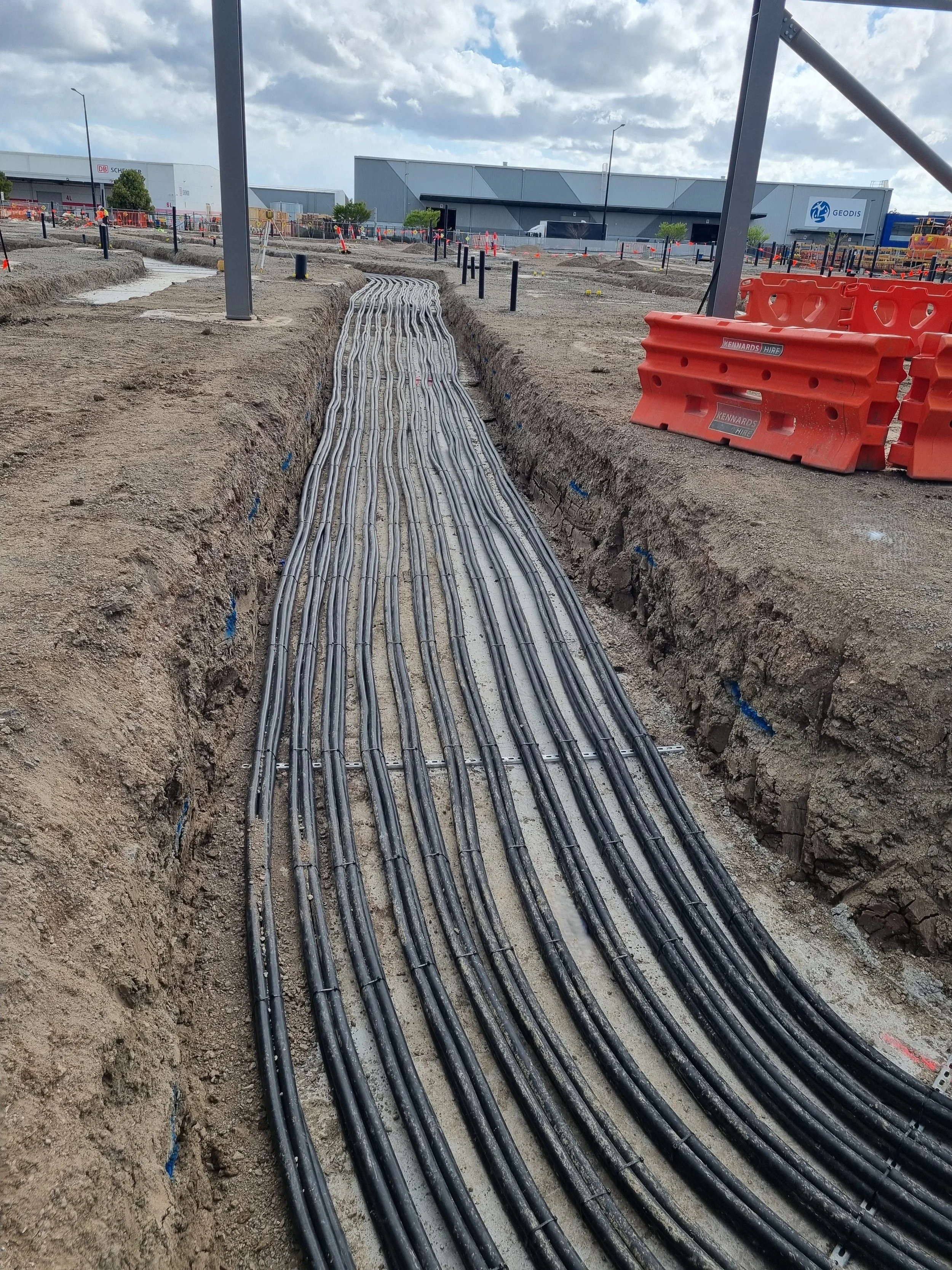 Construction site with multiple electrical cables laid in a trench, orange safety barriers, and industrial buildings in the background under a cloudy sky.