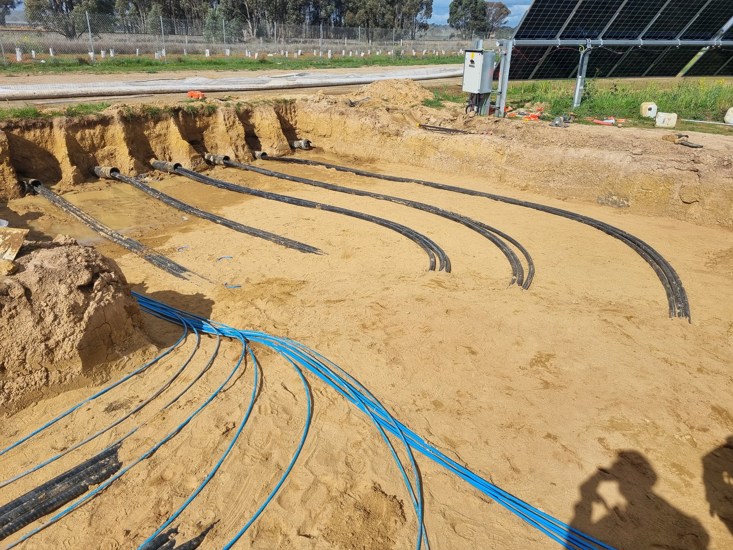 Under construction site with multiple black and blue cables running into underground conduits in sandy dirt, with solar panels and equipment visible in the background.