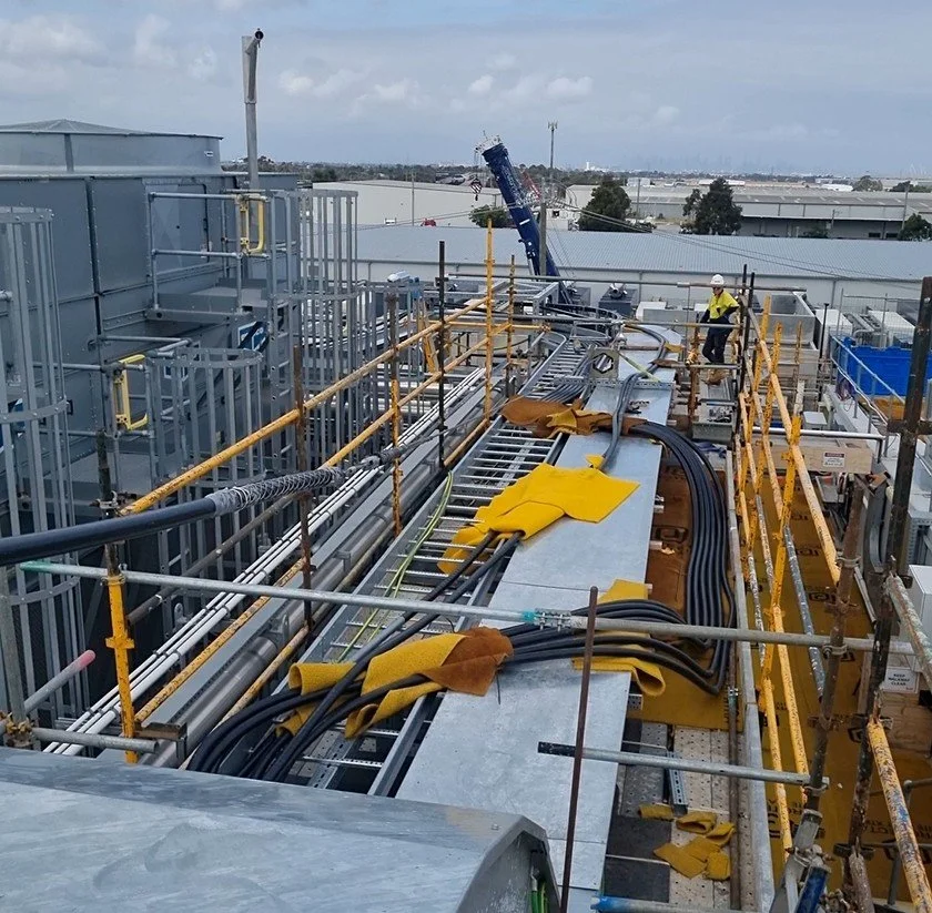 Construction workers working on elevated platform with safety railings, surrounded by various cables, pipes, and construction equipment in an industrial area with warehouses.
