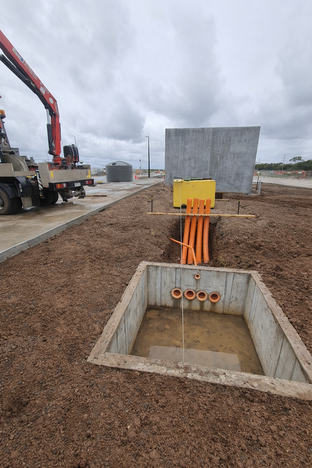 Construction site with an open trench filled with water, orange conduits leading to a large concrete utility box, and a yellow electrical box. There is a grey wall or barrier and a red crane truck on the left, with cloudy weather overhead.