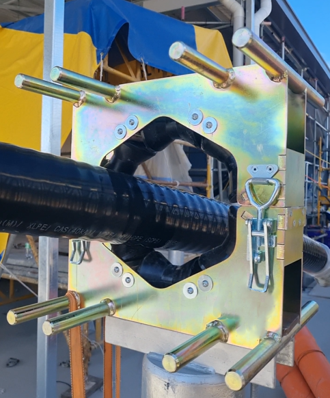 Close-up of a hydraulic pipe bushing with a metal frame and lifting eye, attached to a construction or industrial site with scaffolding and yellow and blue tarps in the background.