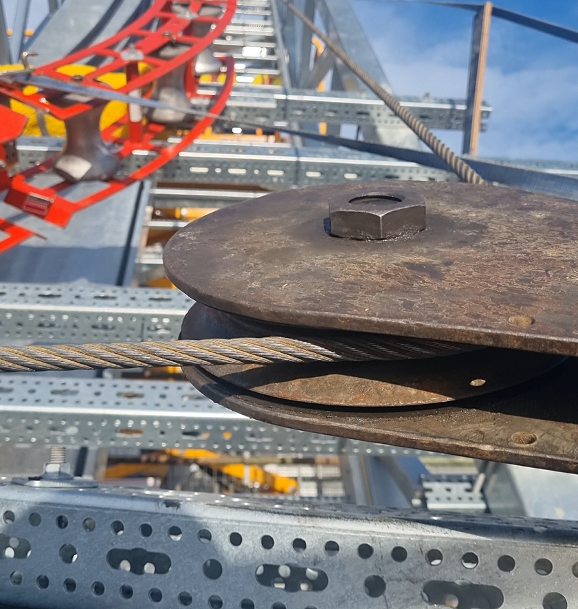 Close-up of a pulley wheel with a steel cable passing through it, the pulley is mounted in a cable tray on a steel structure.