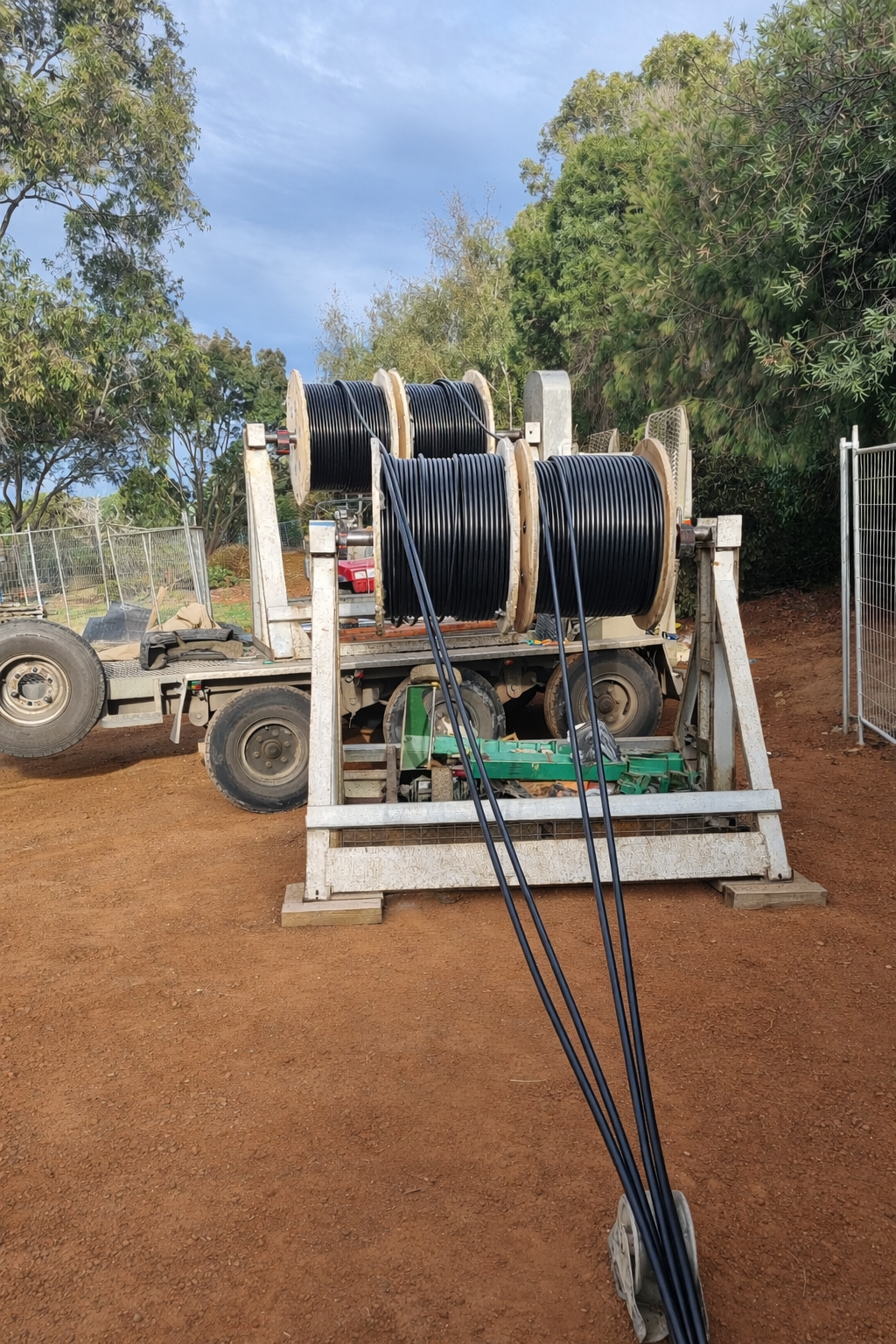 A flatbed truck with large spools of black electrical cable mounted on it, set outdoors on a dirt ground surrounded by trees and a metal fence in the background.