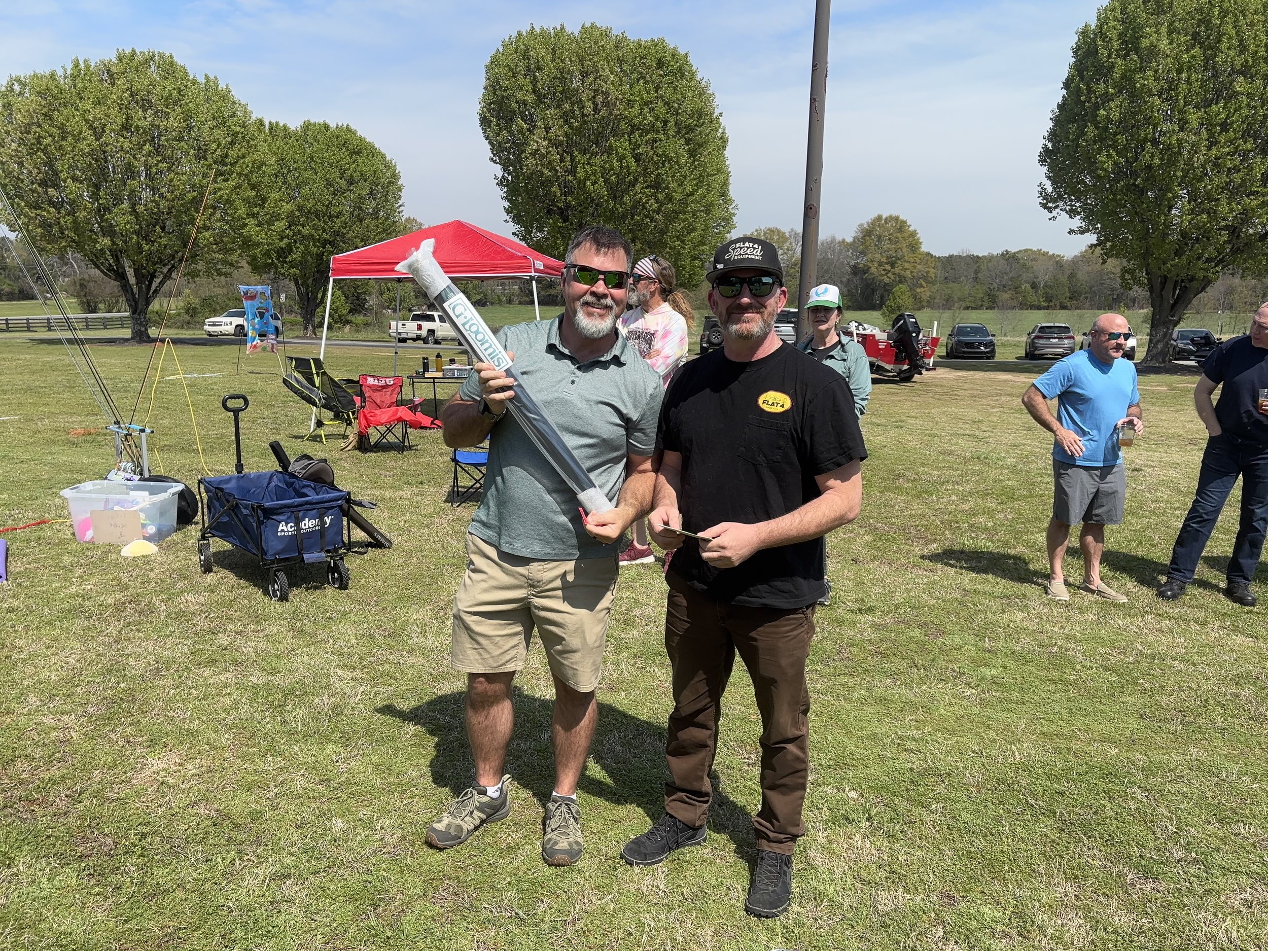 Two men standing on a grassy field at an outdoor event, one holding a large inflatable object and smiling. Several other people are in the background, some holding drinks near tents and parked cars, with trees and a blue sky above.