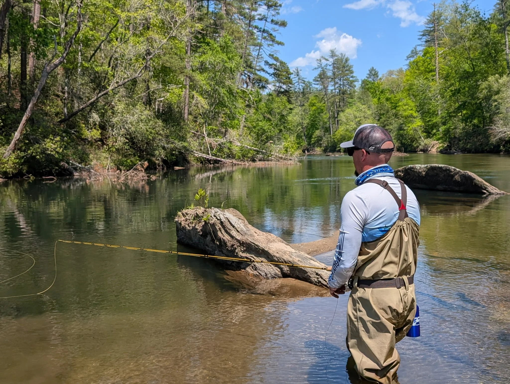 A man standing by a riverbank, wearing a cap, sunglasses, and fishing gear, holding a fishing rod in a forested area with trees, rocks, and a clear blue sky.