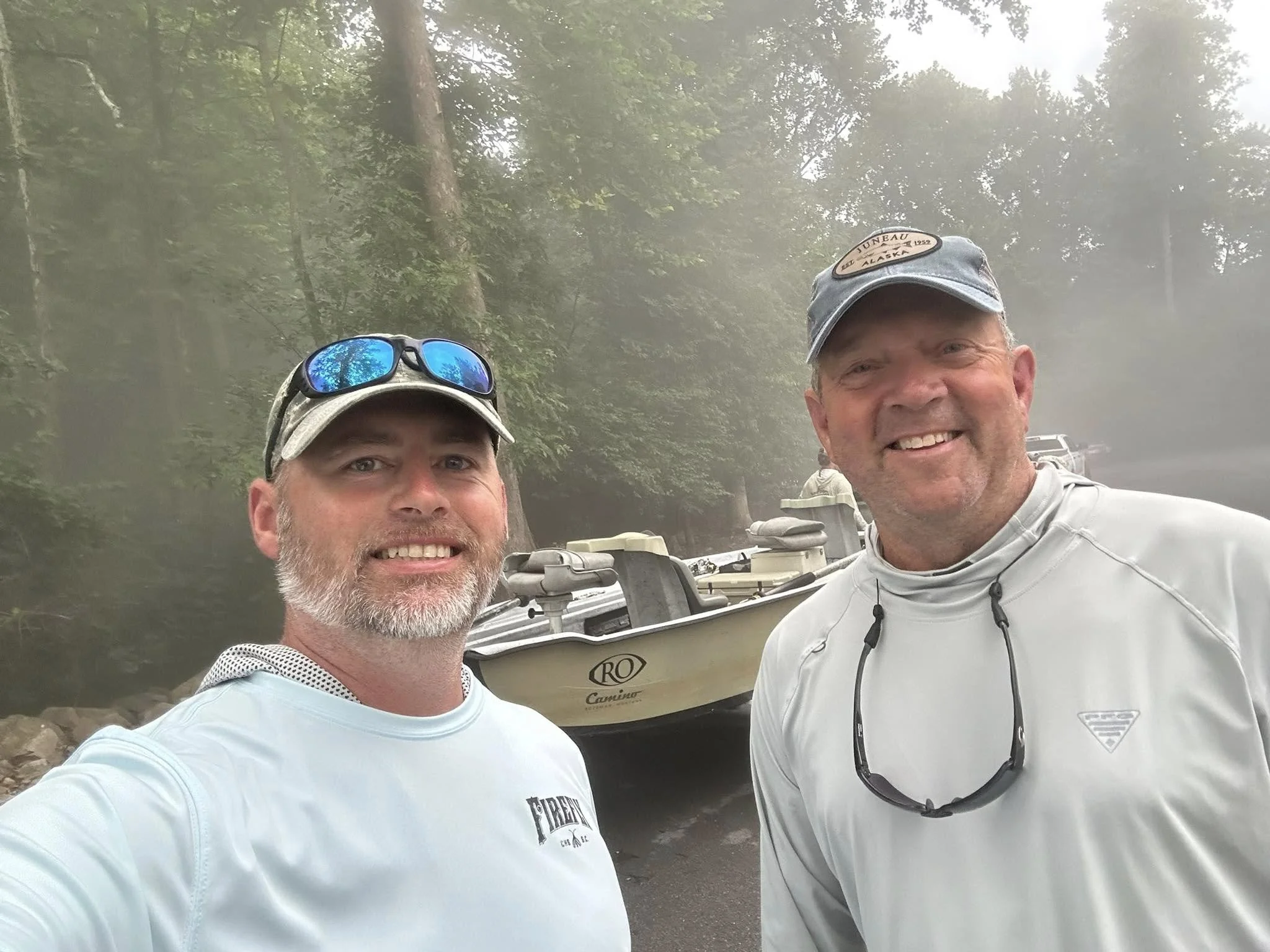 Two men smiling take a selfie outdoors near a boat with a forested background.