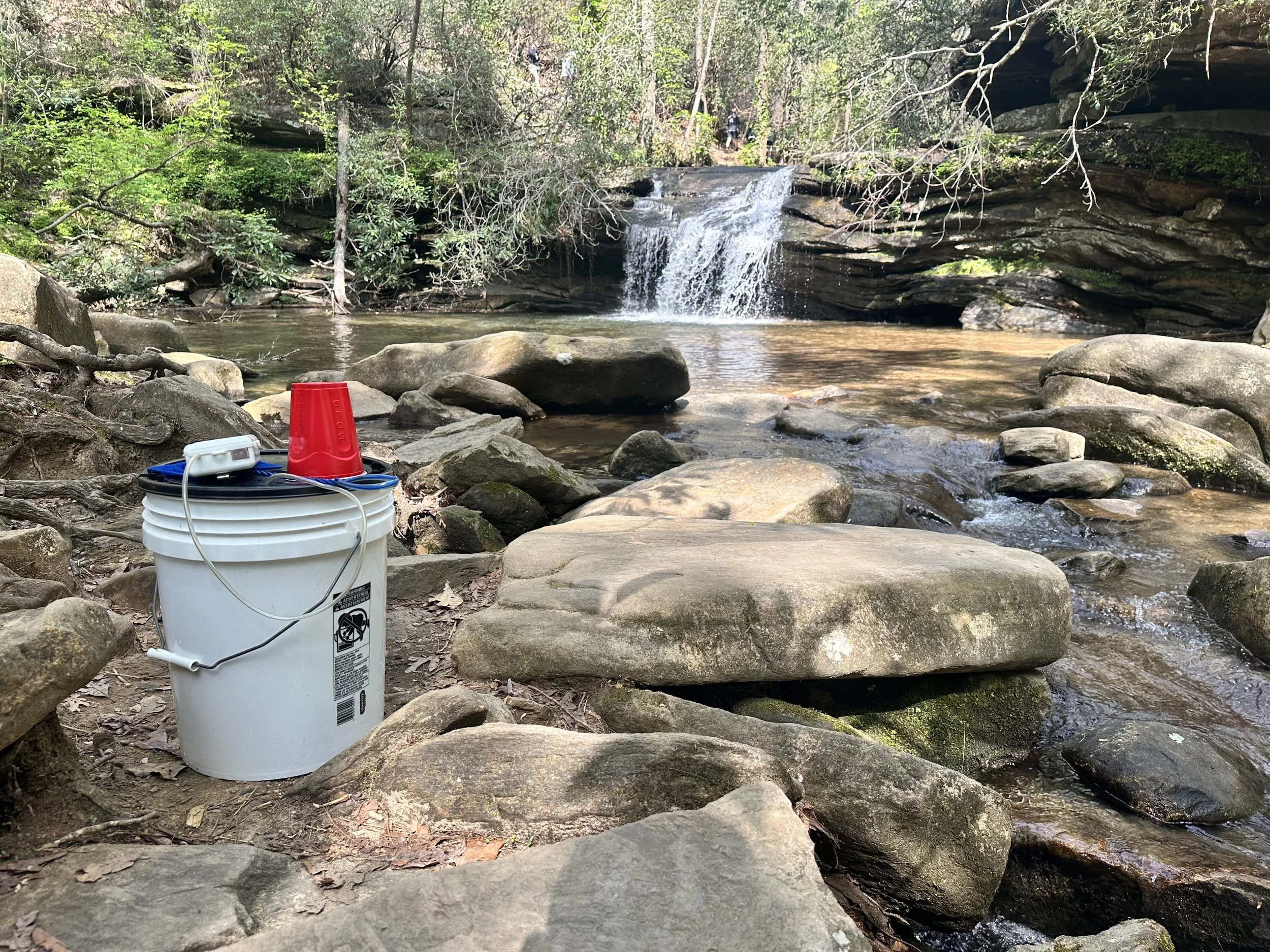 A creek with rocks and a small waterfall in a forest setting, with a white bucket, red cup, and miscellaneous items in the foreground.