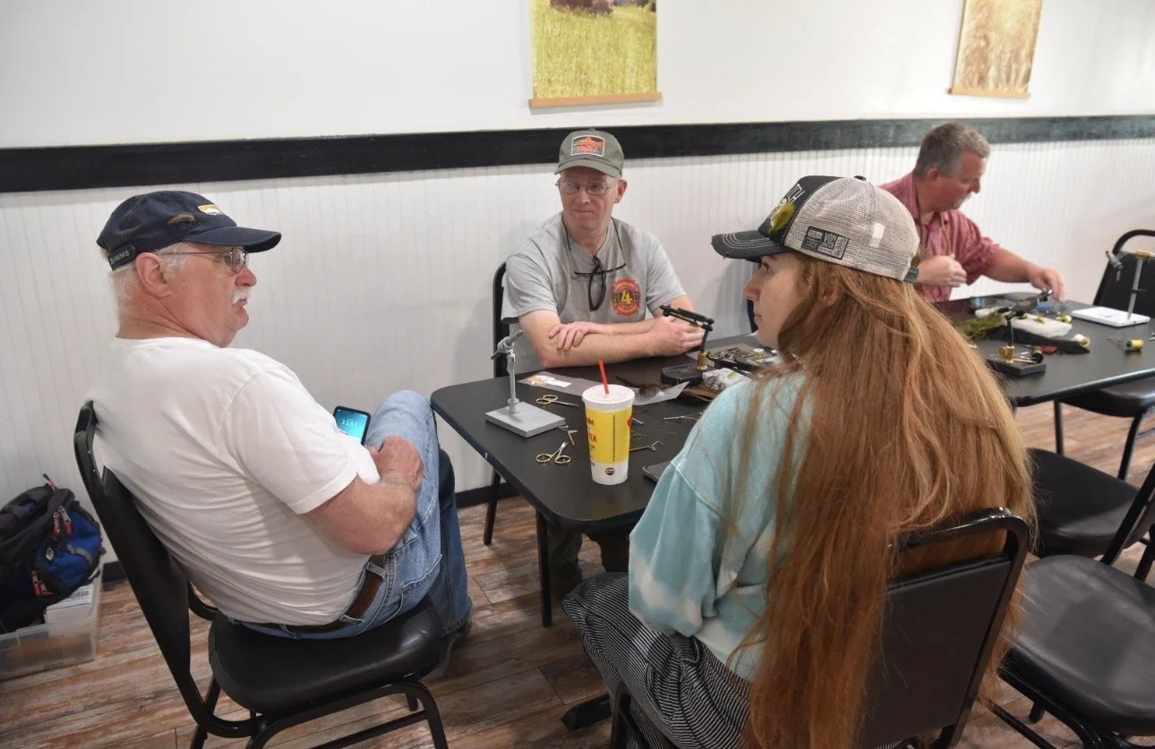 Four people sitting around a table assembling firearms in a room with framed pictures on the wall.
