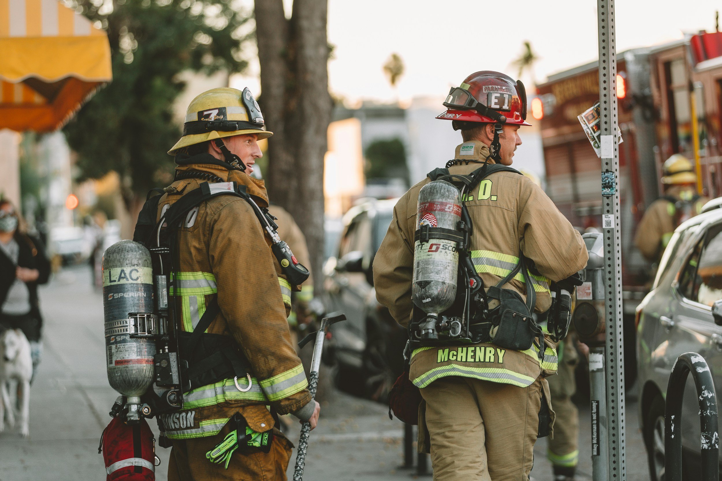 Two firefighters in gear and helmets stand on a city sidewalk, with one looking towards the street and the other facing a pole in front of them. There are cars, trees, and pedestrians in the background.