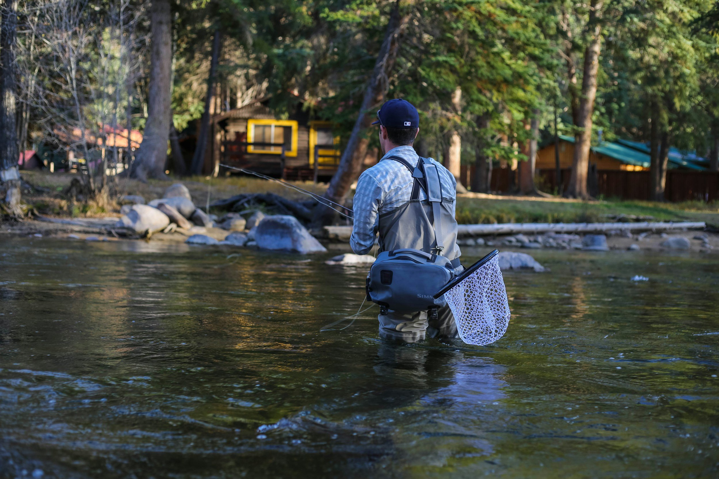 A man fishing in a shallow river with trees and wooden cabins in the background.