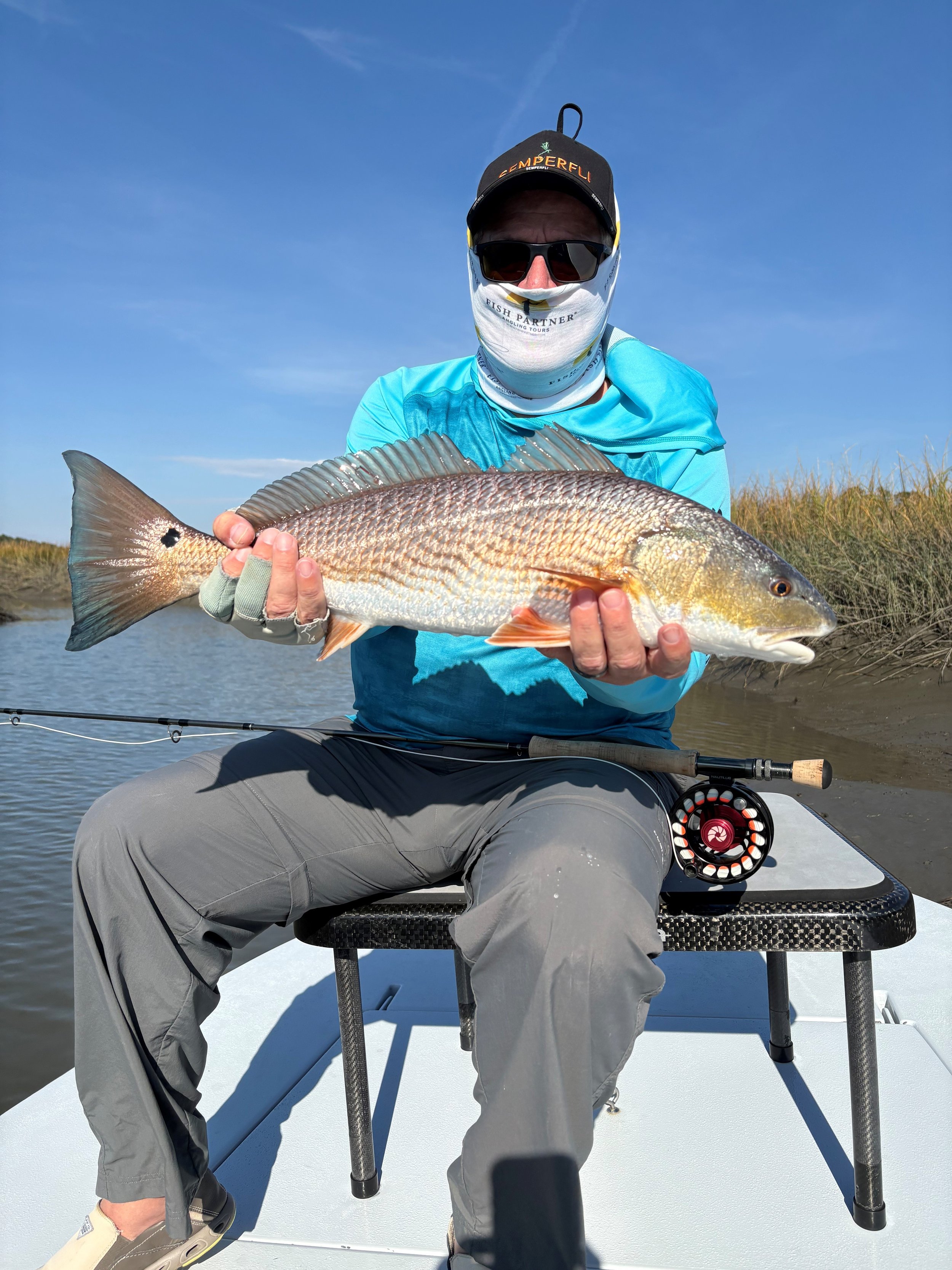 Man sitting on boat holding a large fish with water and marsh grass in the background, wearing sunglasses, cap, and fishing gear.