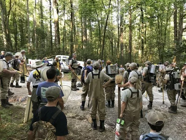 Group of people conducting a forestry or environmental experiment in a wooded area, with some equipment and vehicles in the background.