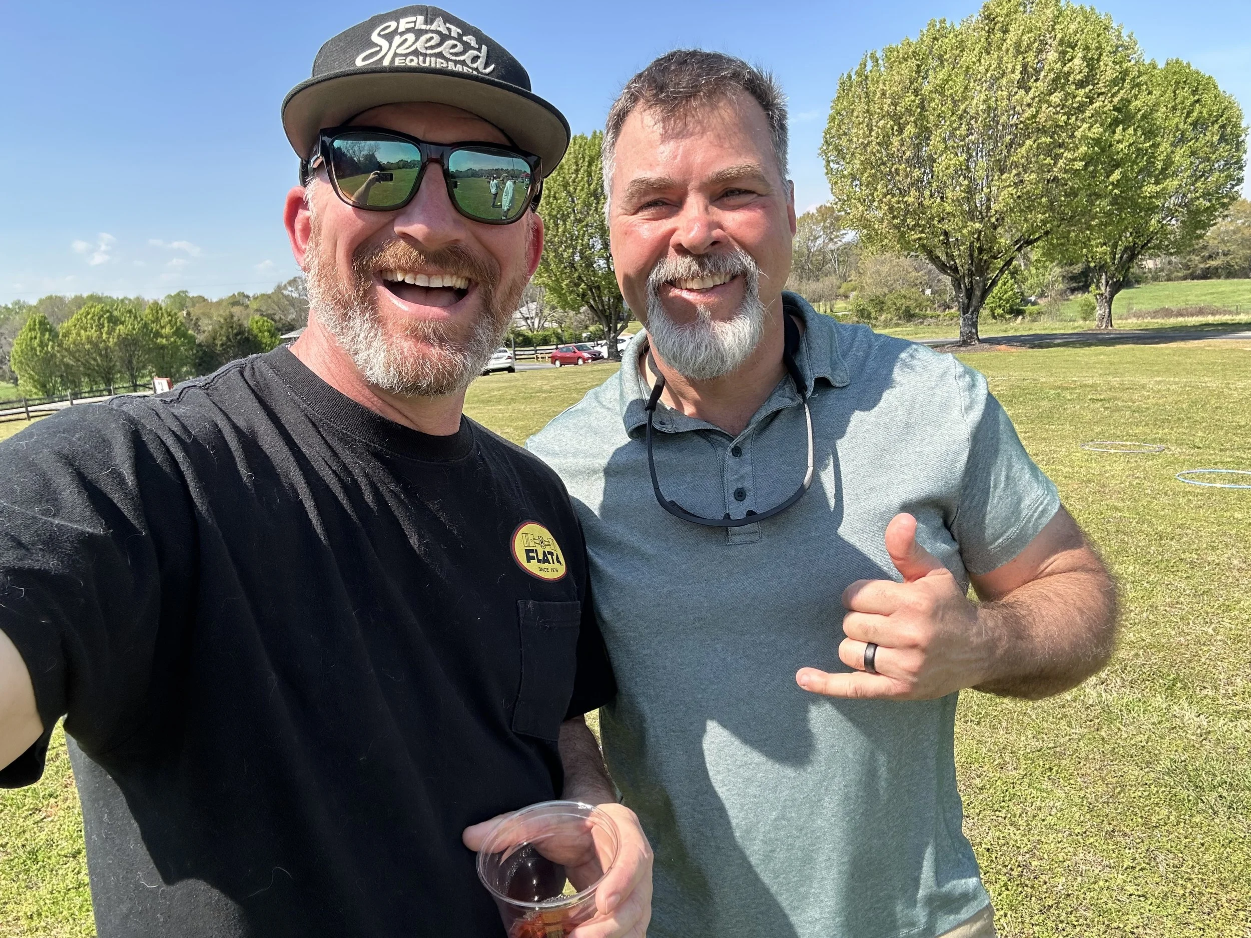 Two smiling men outdoors on a sunny day, one wearing sunglasses and a baseball cap, the other with sunglasses hanging from his collar, standing in a grassy field with trees and parked cars in the background.