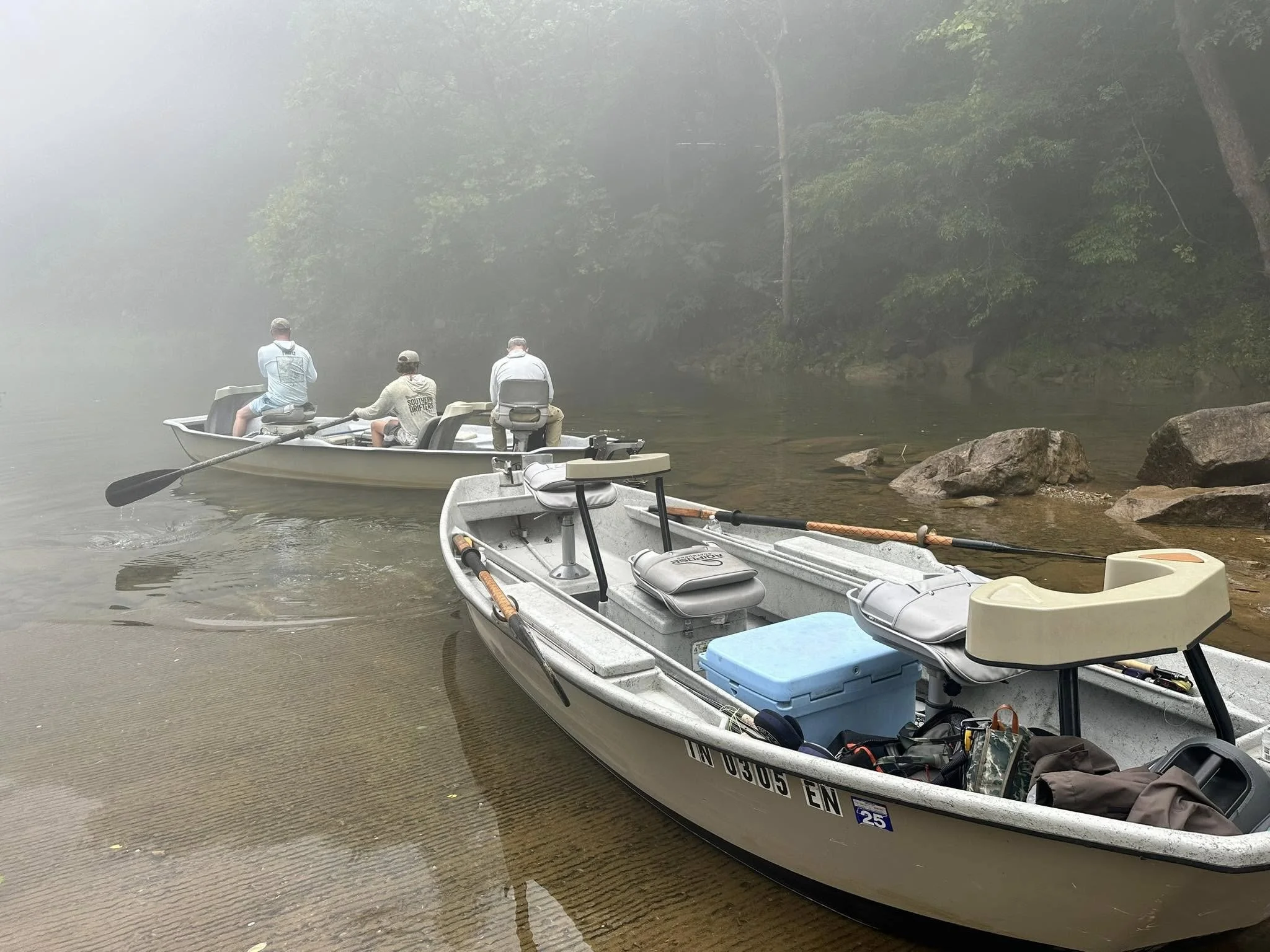 Three men fishing on a foggy river with two boats and rocks along the shoreline.