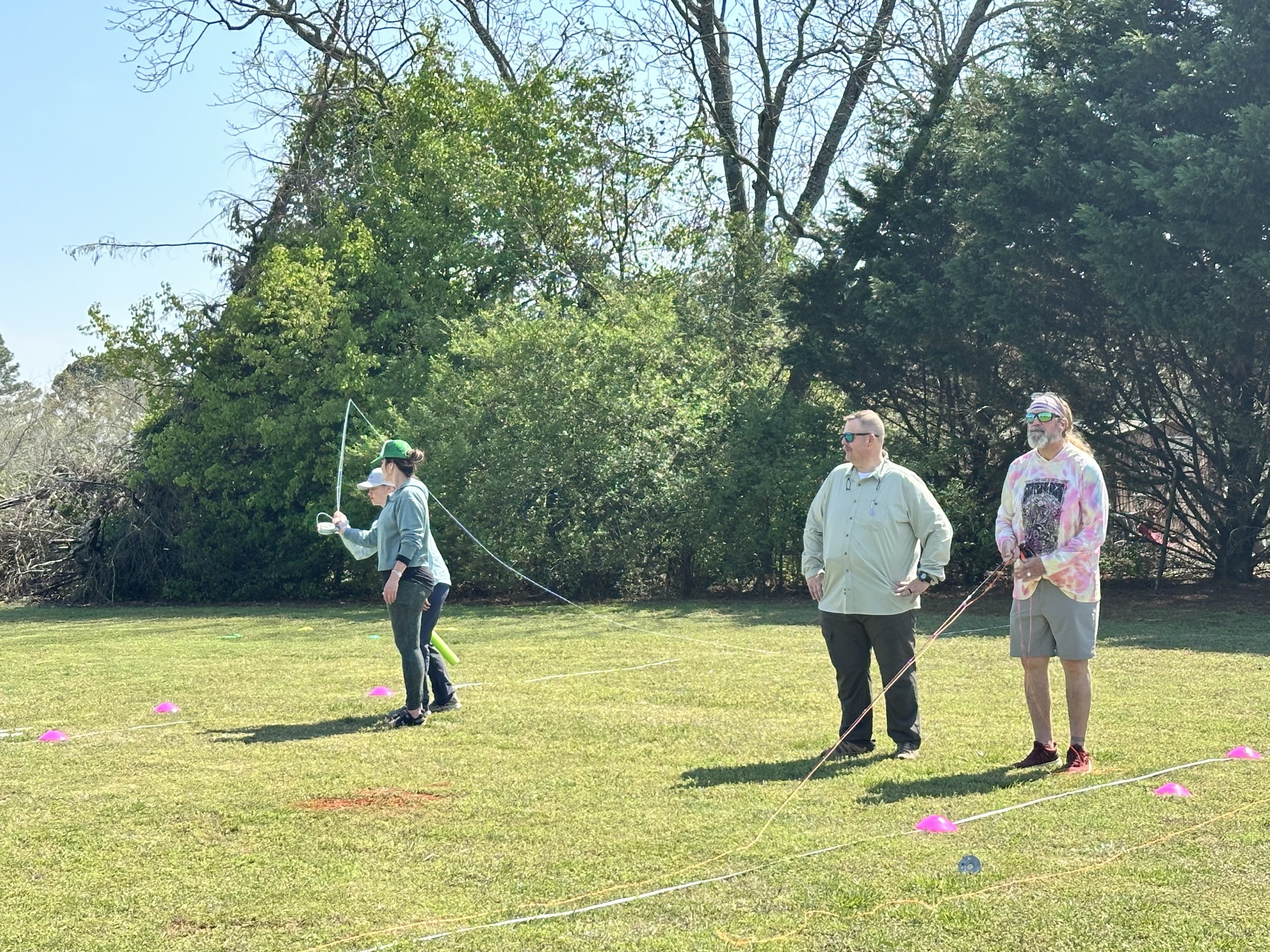 People standing on grass field with trees in the background, some holding equipment and others observing, possibly participating in outdoor activity or workshop.