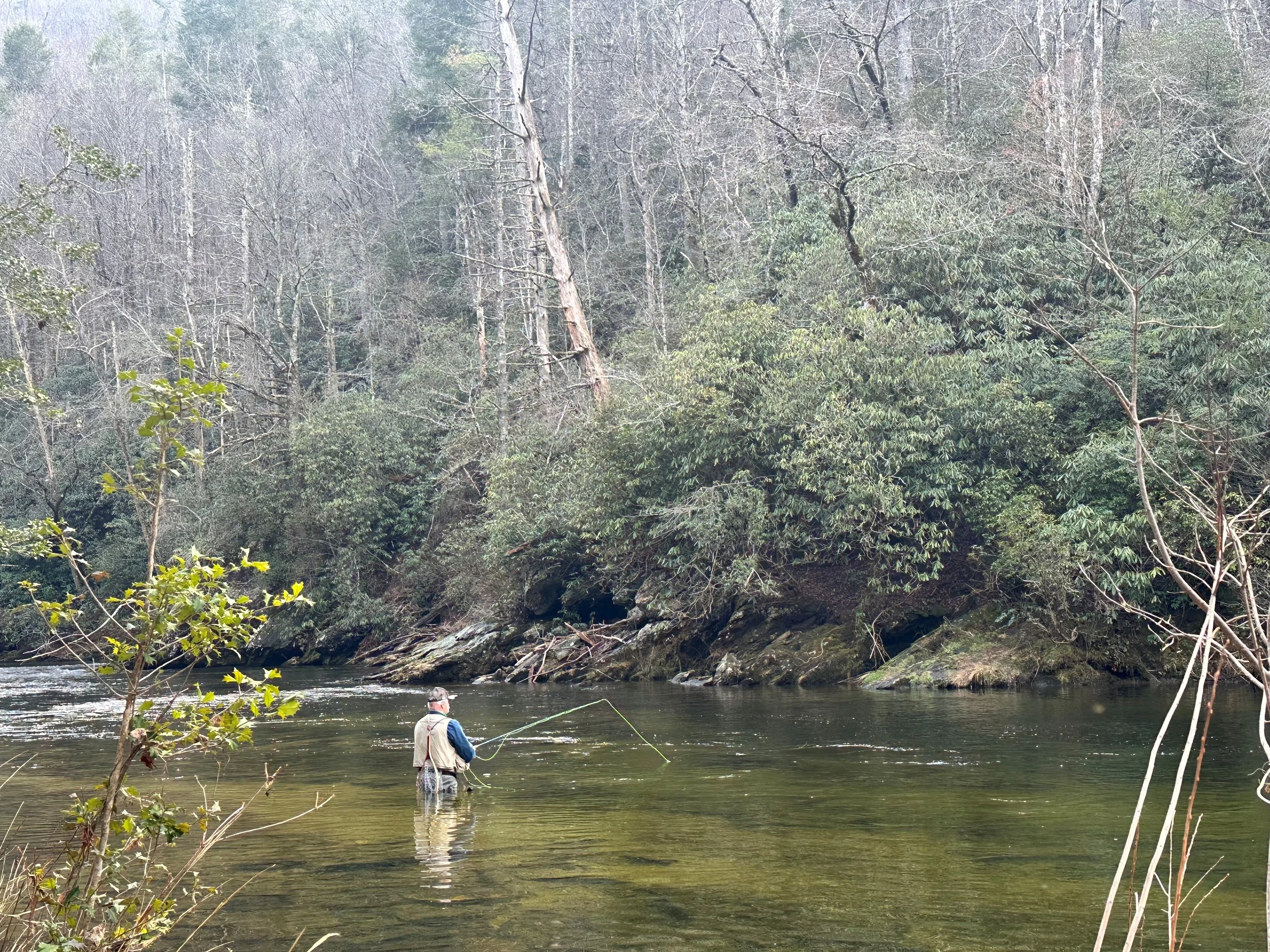Man fishing in a river surrounded by dense trees and rocks on the riverbank.