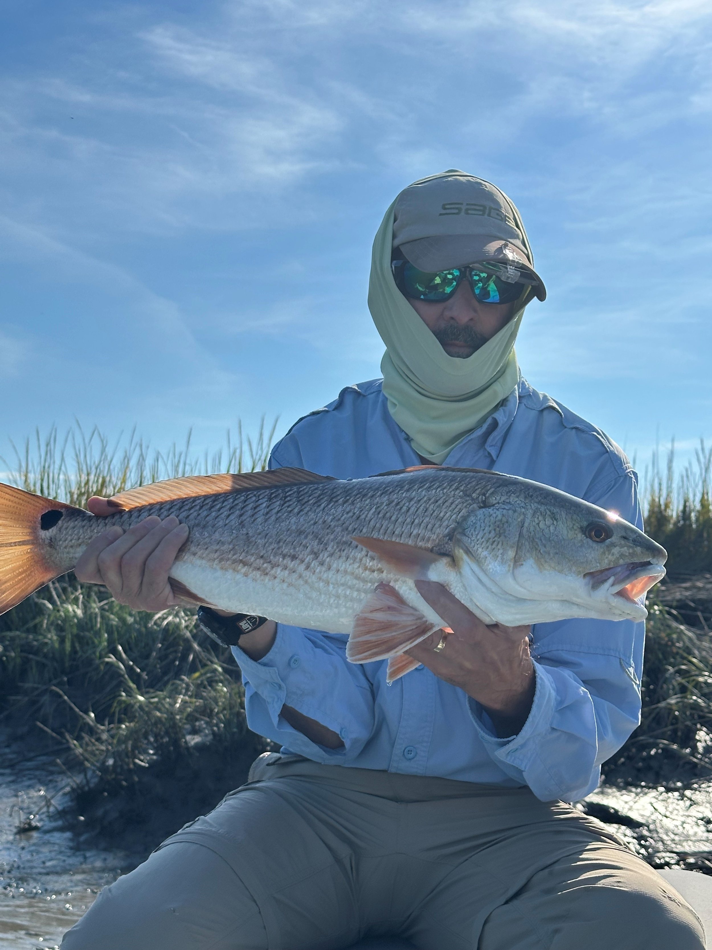 Man in sun-protective gear holding large fish outdoors with blue sky and grassy background.