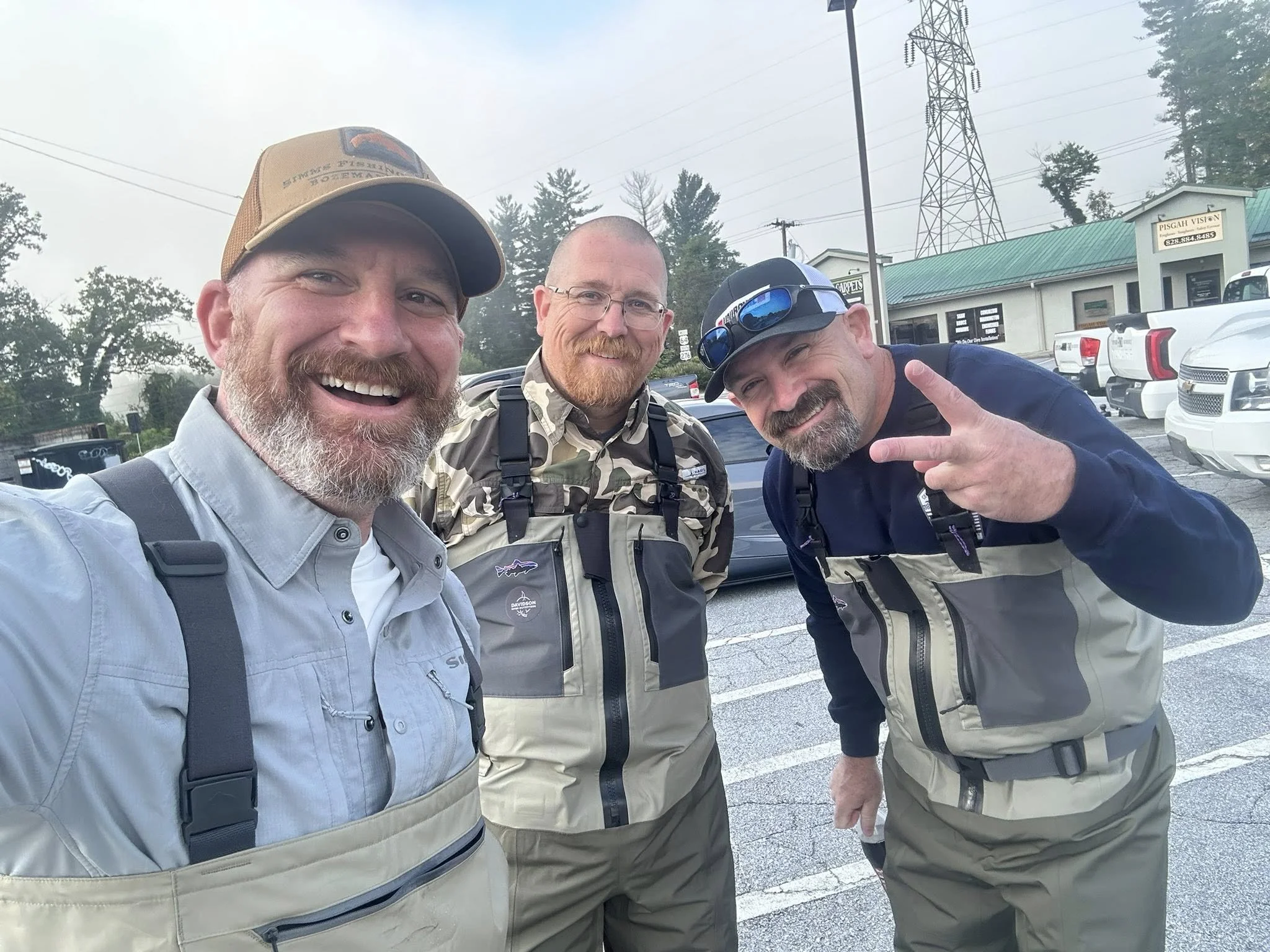 Three men in fishing gear taking a selfie in a parking lot, smiling and making a peace sign, with trees and a building in the background
