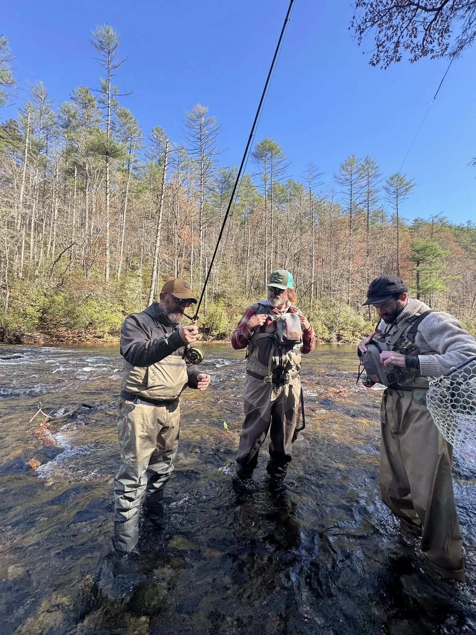 Three men fishing in a shallow river, surrounded by trees with a clear blue sky overhead.