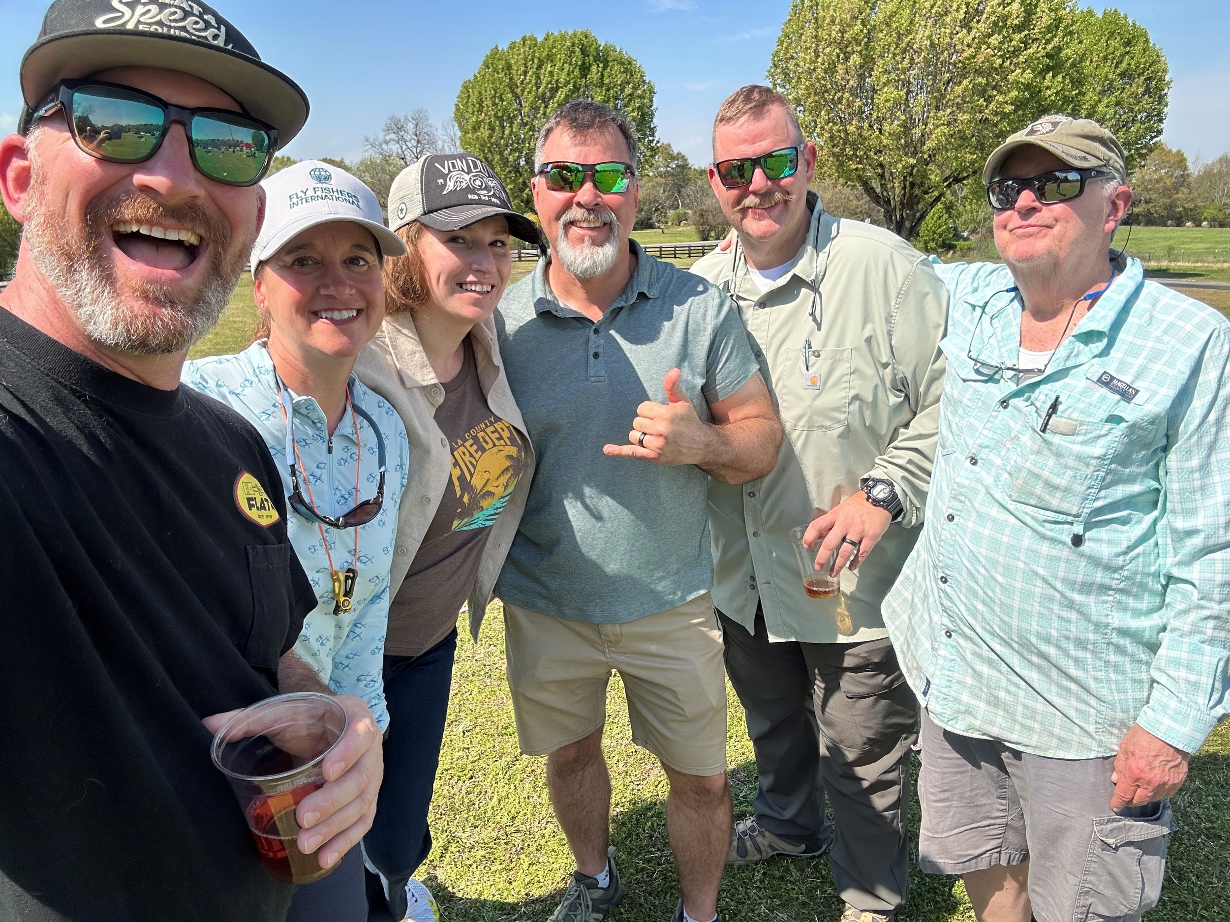 Group of six people standing outdoors on a sunny day, smiling. They are wearing casual clothing and sunglasses, with two people holding drinks. Trees and a fence are visible in the background.