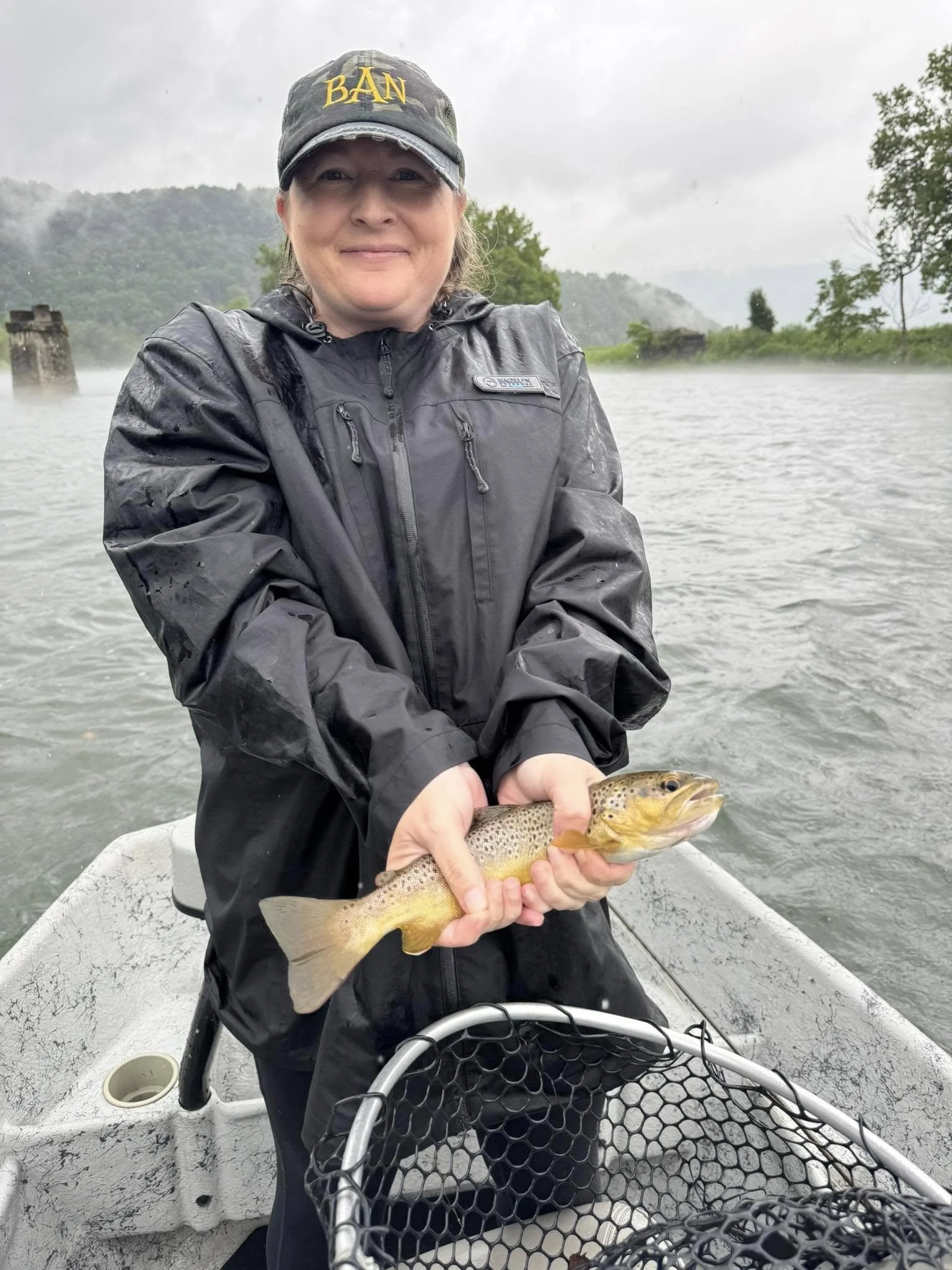 Woman in black jacket and cap holding a fish on a boat, with river and green trees in the background on a rainy day.