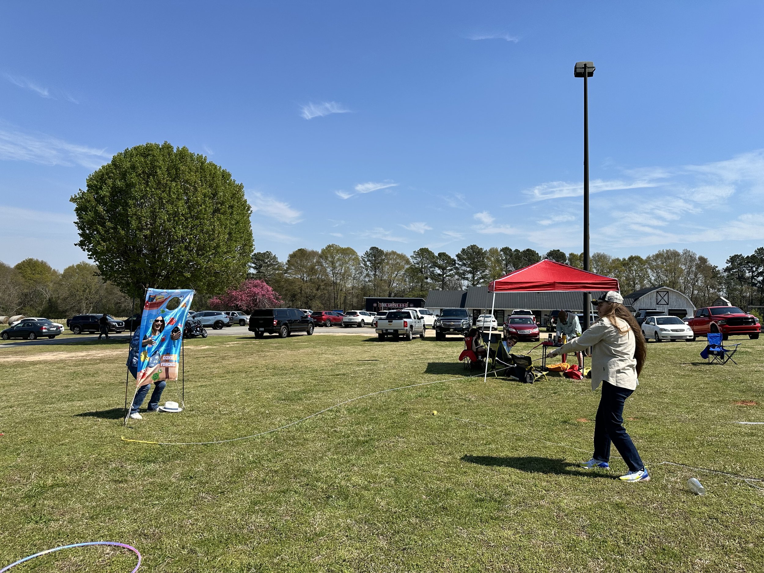 People playing a game with a long string in a park on a sunny day, with parked cars and trees in the background.
