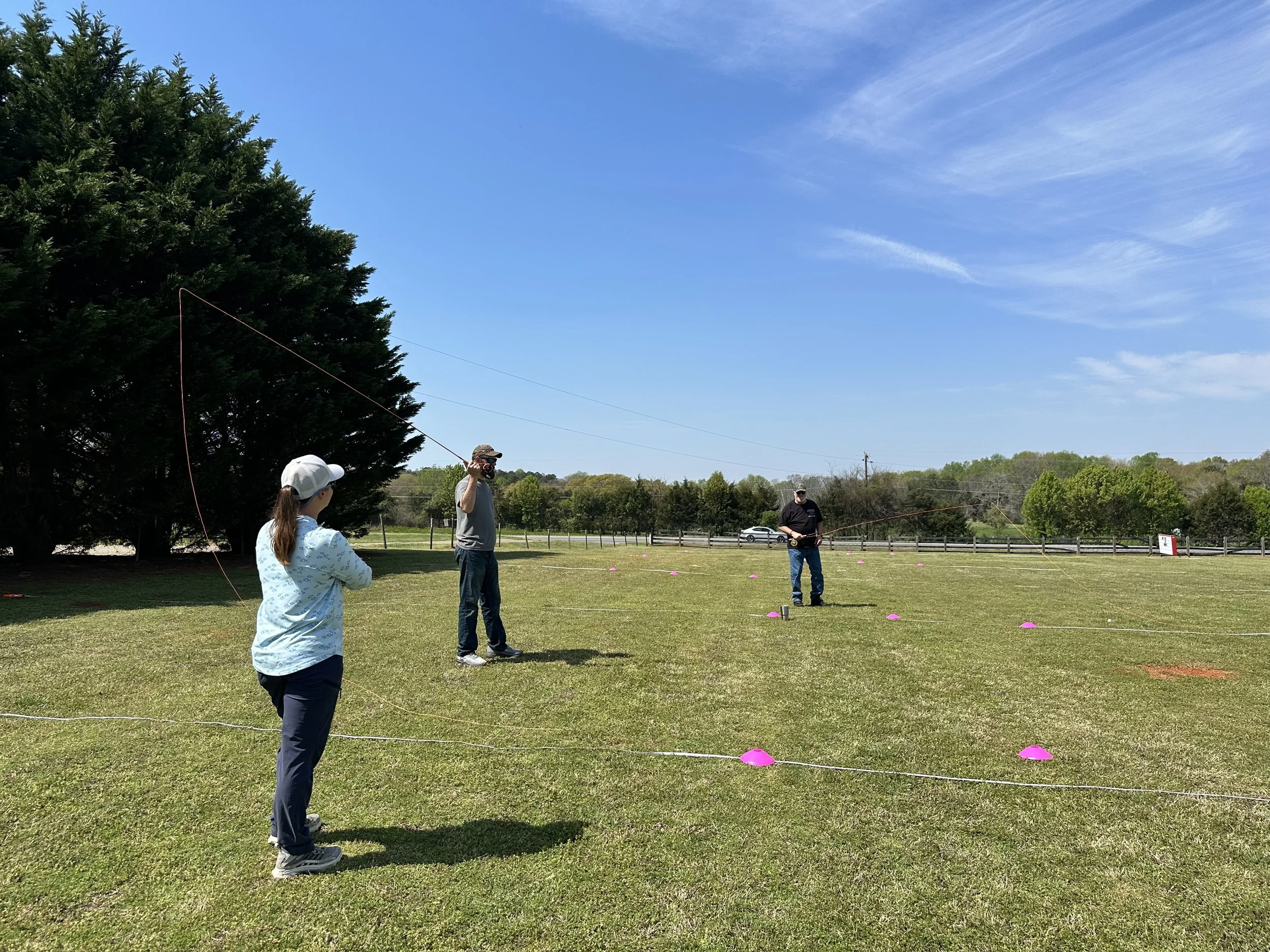 Three people outdoors on a grassy field, flying kites on a clear, sunny day. One woman is facing away from the camera with a baseball cap, two men are facing each other holding kite strings, and pink cones mark the field.