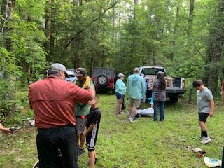 Group of people gathered in a forest clearing near parked vehicles.