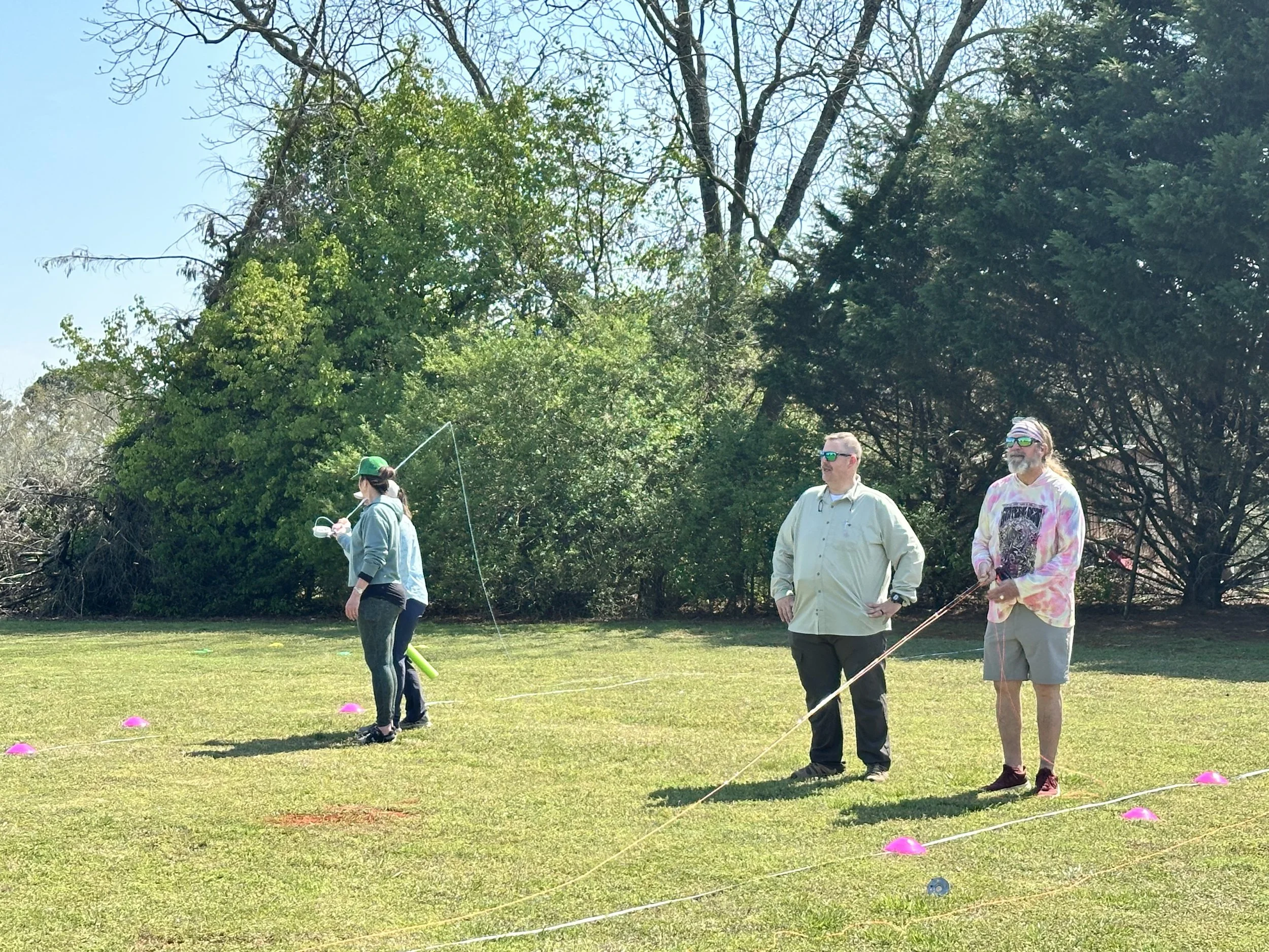 Three people outdoors on a grassy field with trees in the background, two men standing and watching, and a woman in a blue jacket and green cap holding a fishing rod or similar object.
