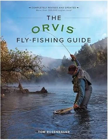 A person fly-fishing in a river, standing knee-deep in water, with a fishing rod in hand, surrounded by trees and mountains.