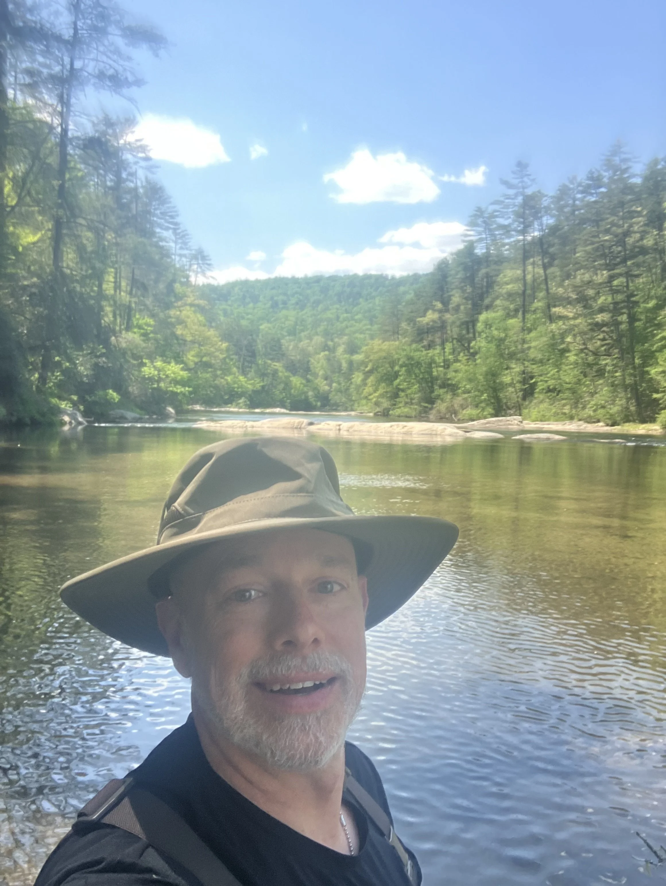A man wearing a large sun hat and sunglasses is standing by a river with a forested hillside in the background under a bright blue sky with a few clouds.