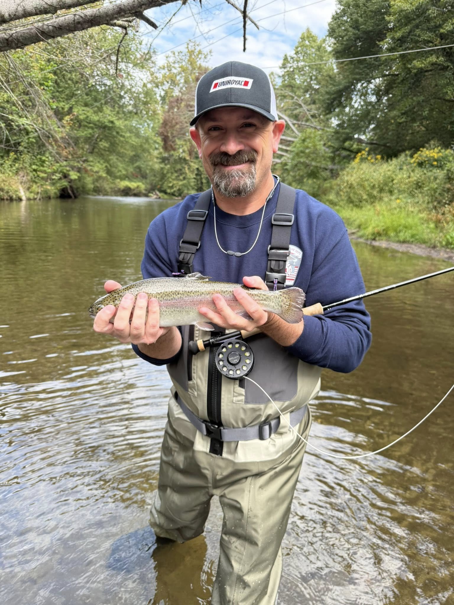 A man standing in a shallow river holding a freshly caught fish, smiling, with trees and blue sky in the background.