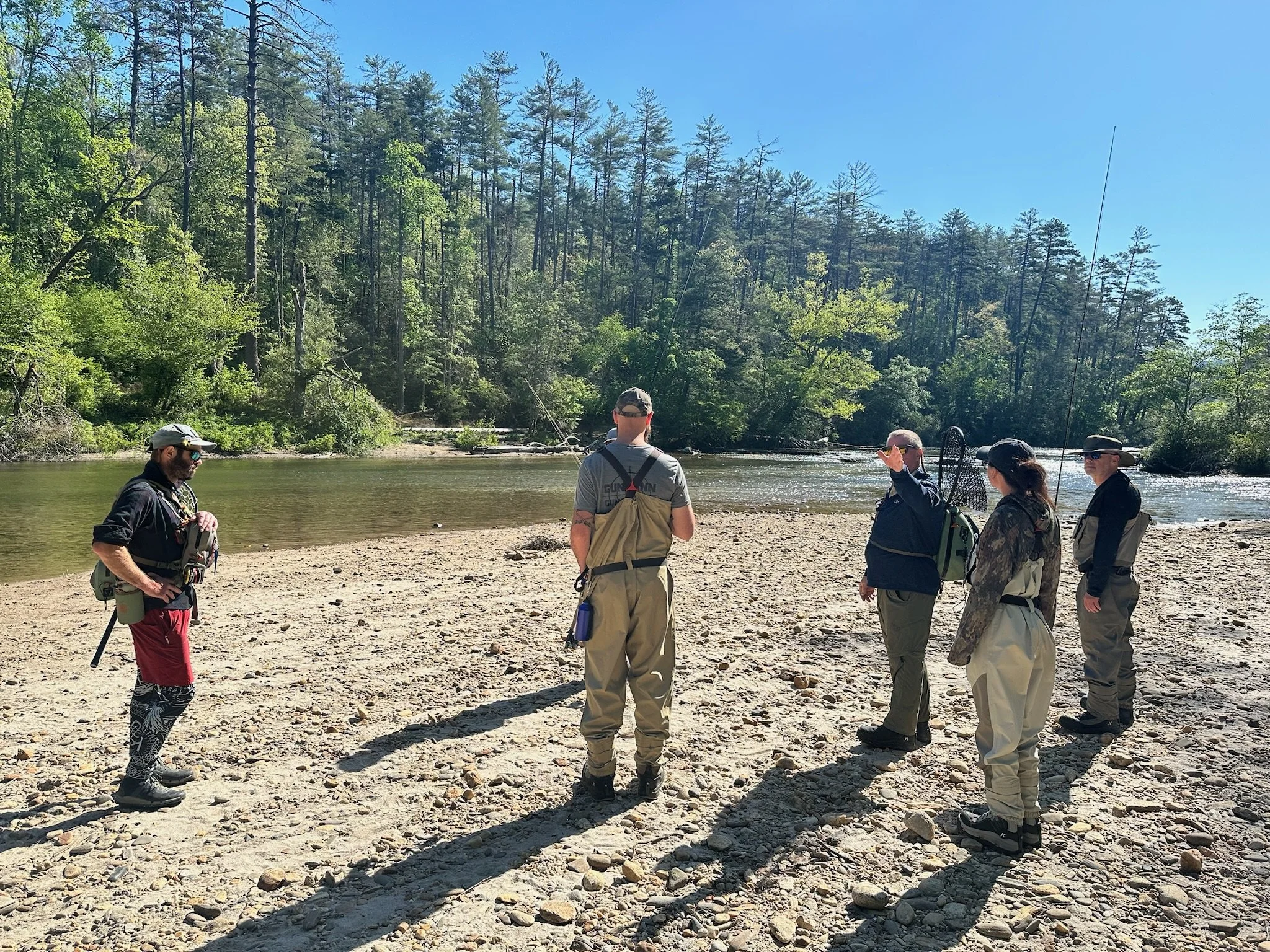 A group of five people standing on a rocky riverbank with fishing gear, engaged in a conversation near a river flowing through a forested area with tall trees and a clear blue sky.