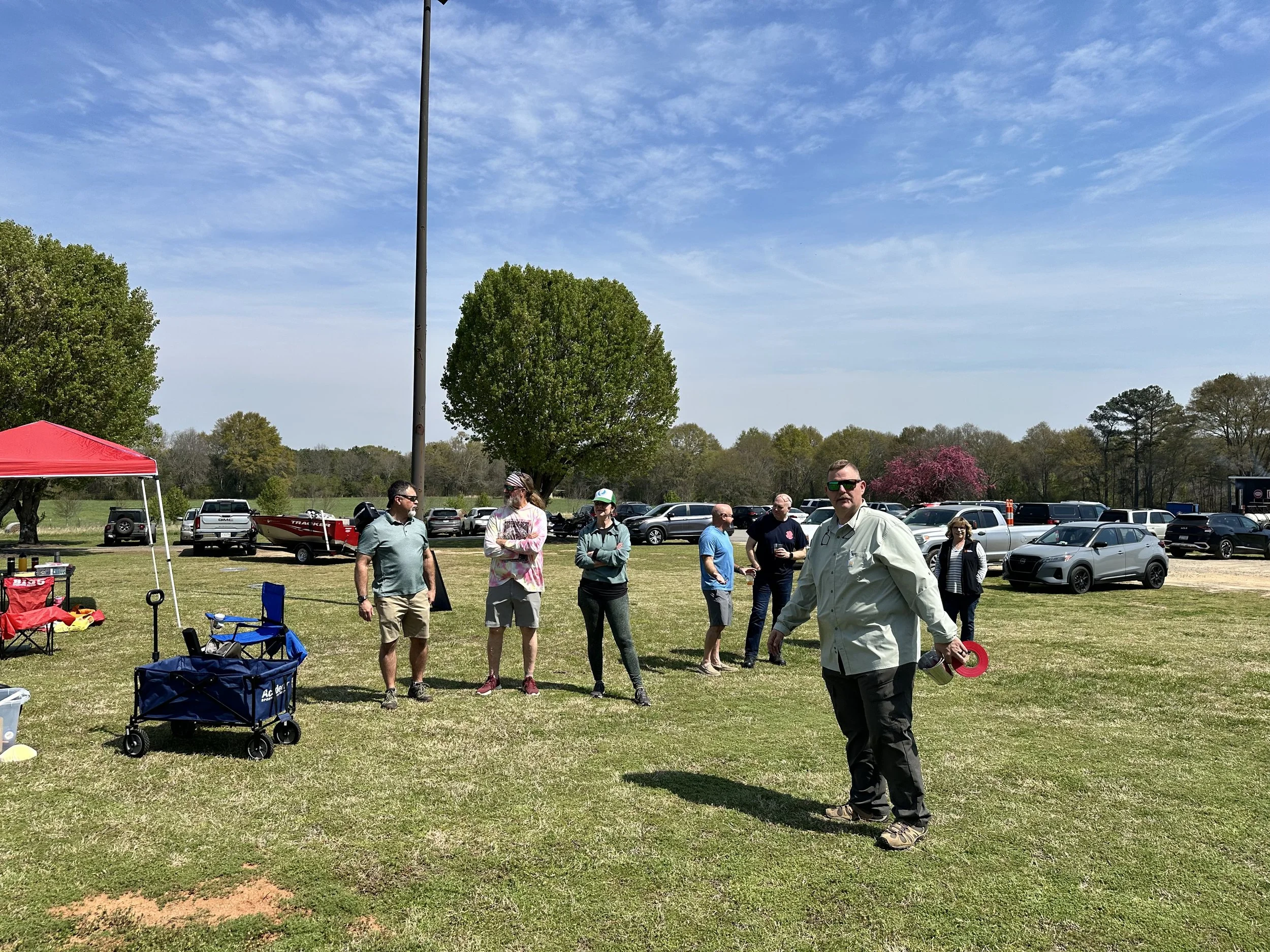 People standing on grass field in a park during daytime, some near a red tent, with trees, parked cars, and a blue sky in the background.